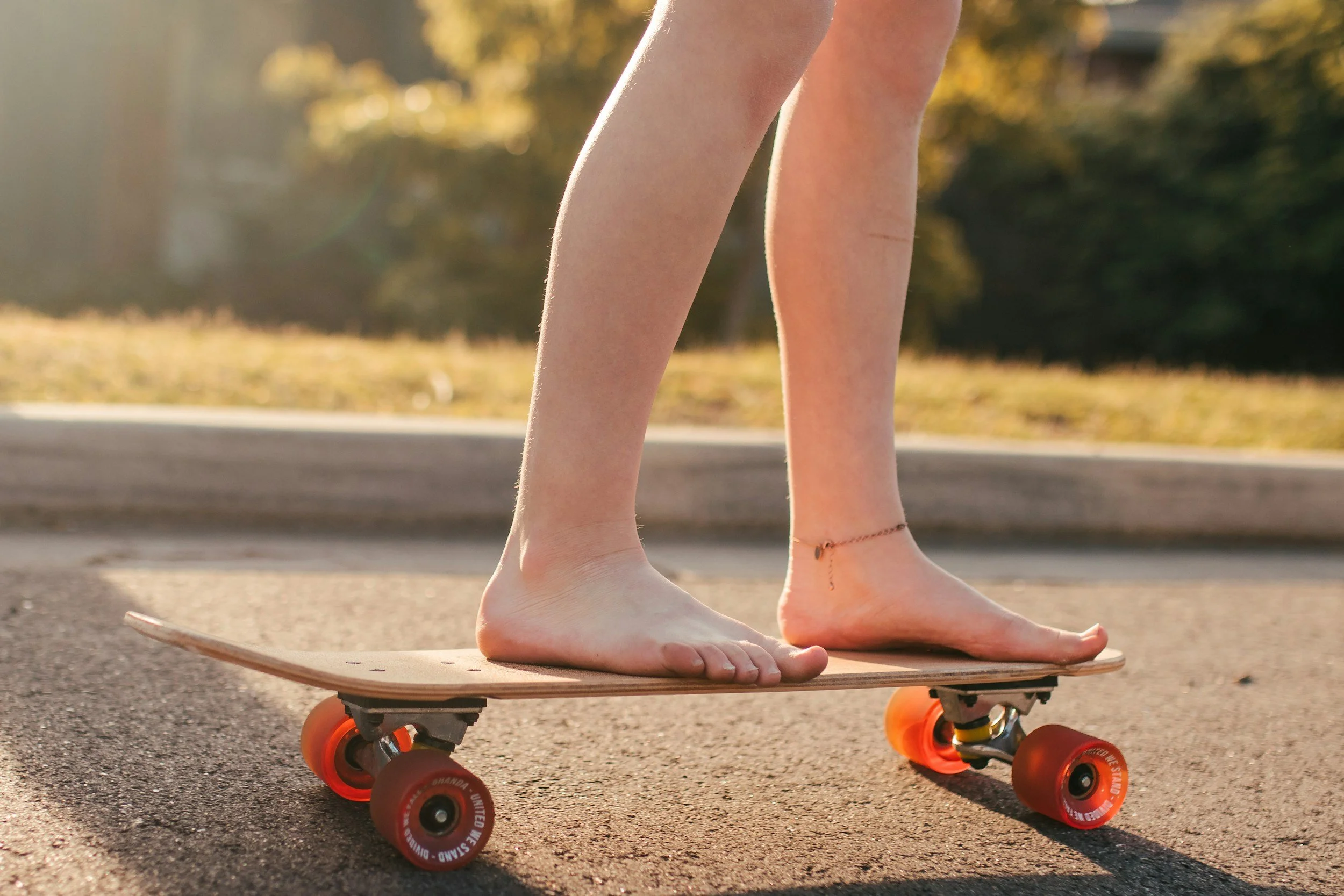 Person barefoot riding a skateboard on pavement in park during sunset, wearing a bracelet around ankle.