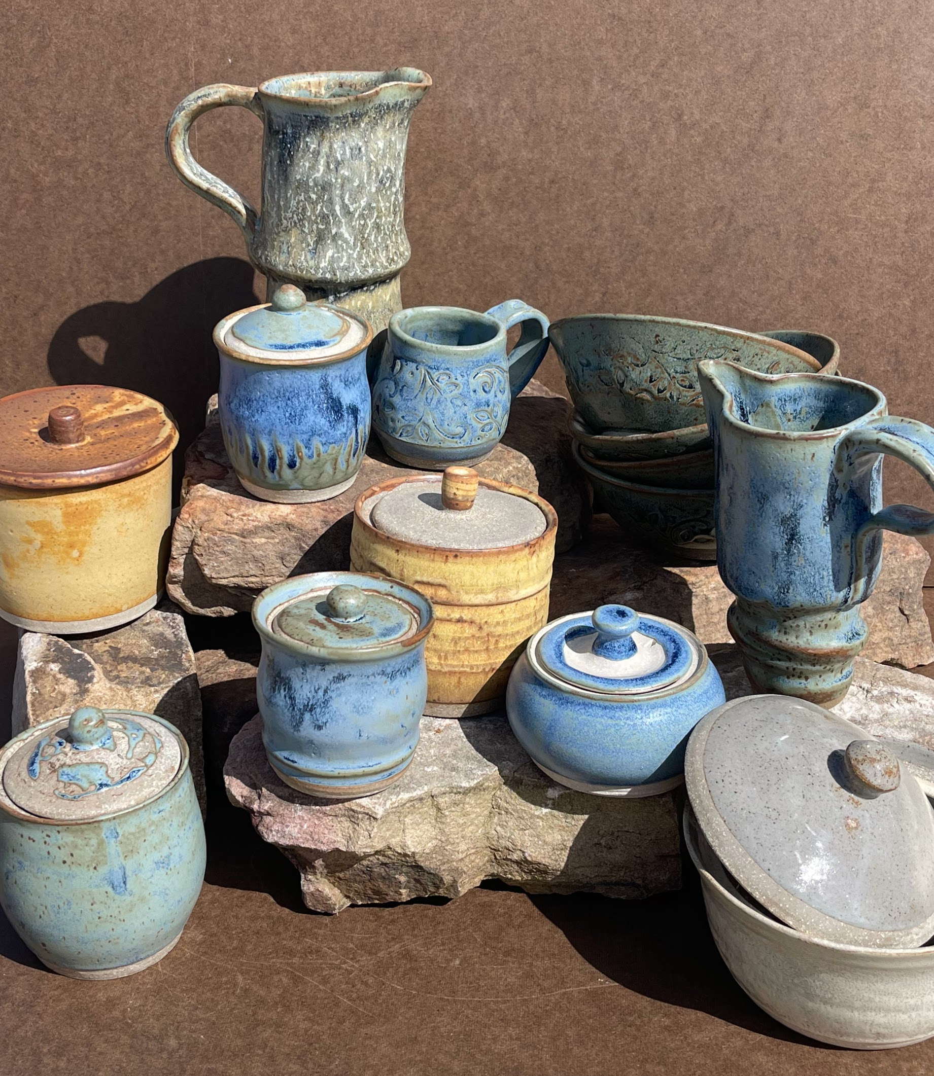 Collection of handcrafted ceramic pottery including jars, cups, bowls, and pitchers with glazed surfaces in blue, green, and beige tones, displayed on a rough stone surface.