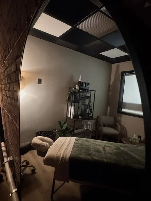 Massage room with a massage table, a black metal shelf with bottles, and a seated chair near a window.