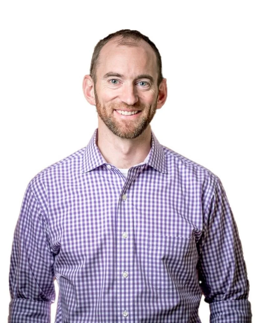 Headshot of a man with short brown hair and a beard, wearing a purple checkered shirt, smiling against a white background.