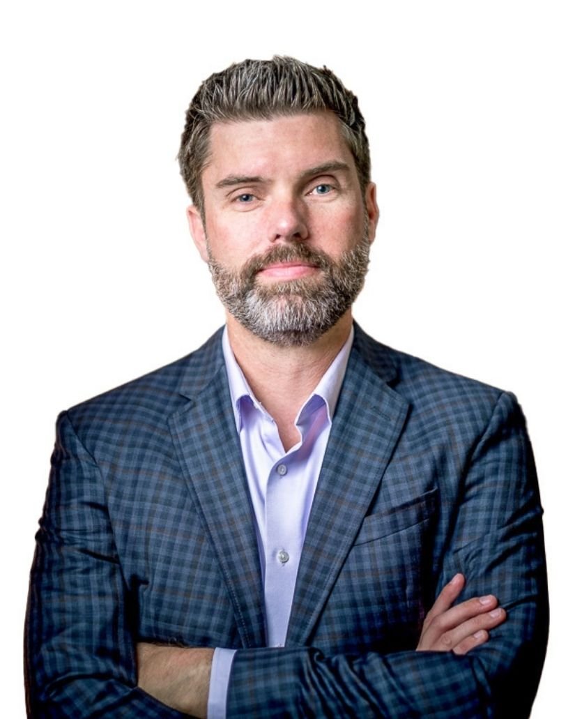 A man with salt and pepper hair and beard, wearing a blue checkered blazer and a light blue collared shirt, standing with arms crossed against a white background.