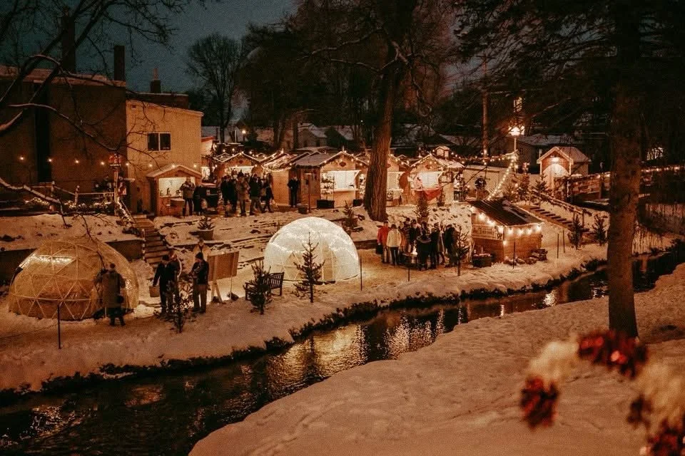 A nighttime scene of a winter holiday market with small decorated stalls, string lights, and people gathered near a small stream.