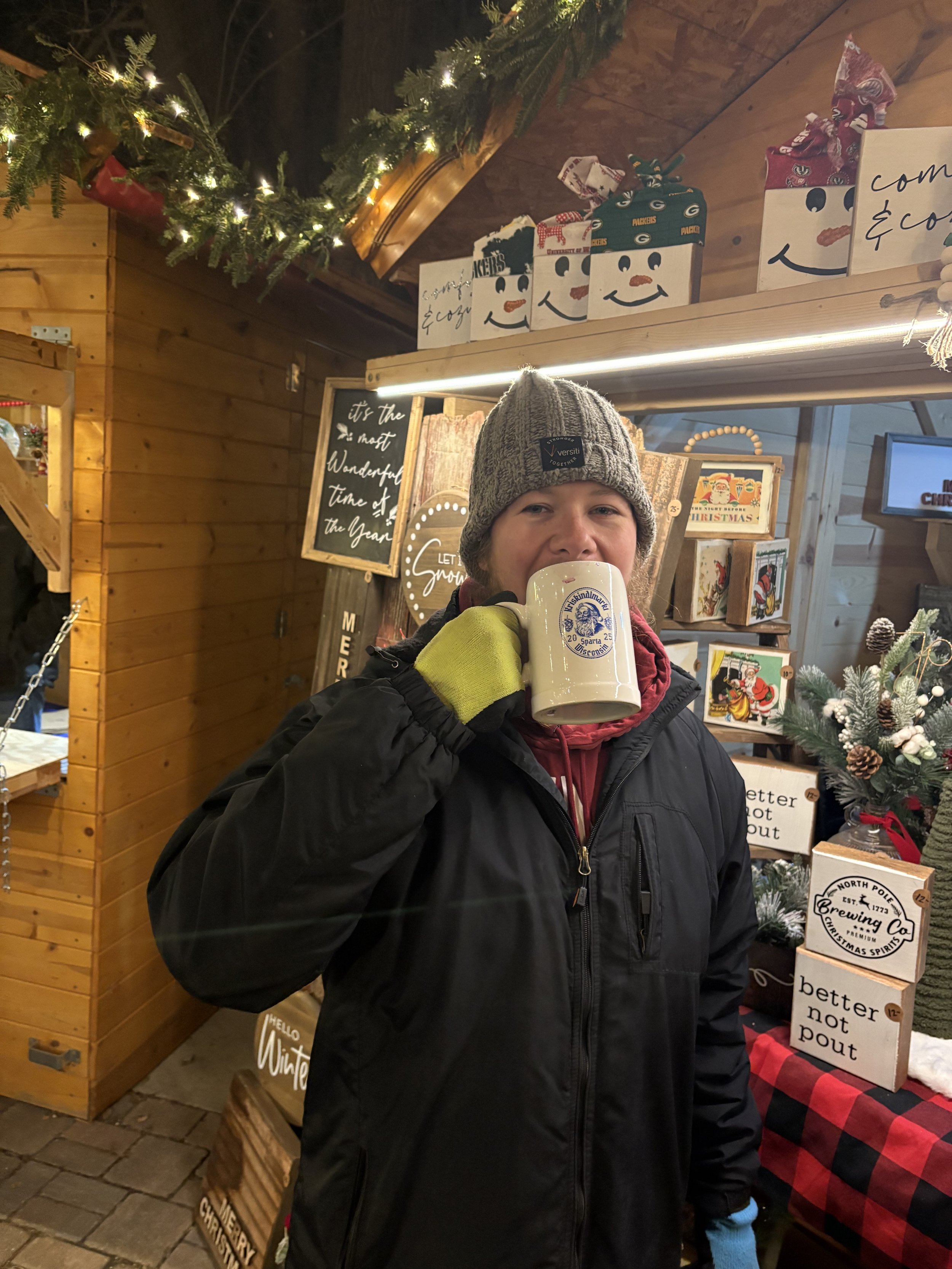 A person wearing a gray knit cap and a black jacket, holding a large beer stein mug, standing at a festive holiday market stall decorated with Christmas items, signs, and wrapped gifts.