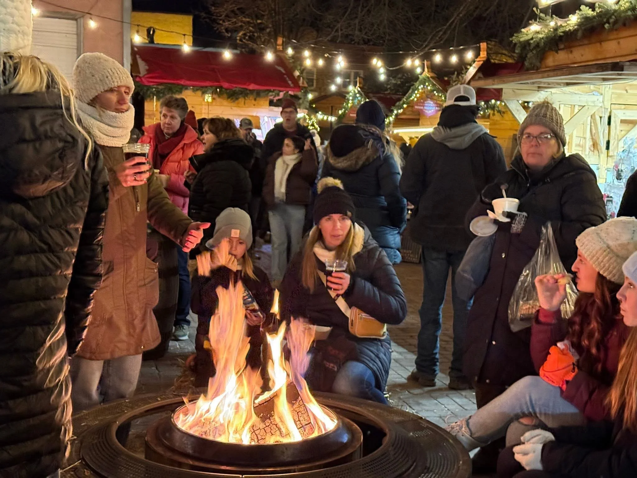 A group of people wearing warm clothing gathered around a fire pit at night, with Christmas lights and decorations hanging above them at an outdoor holiday market.