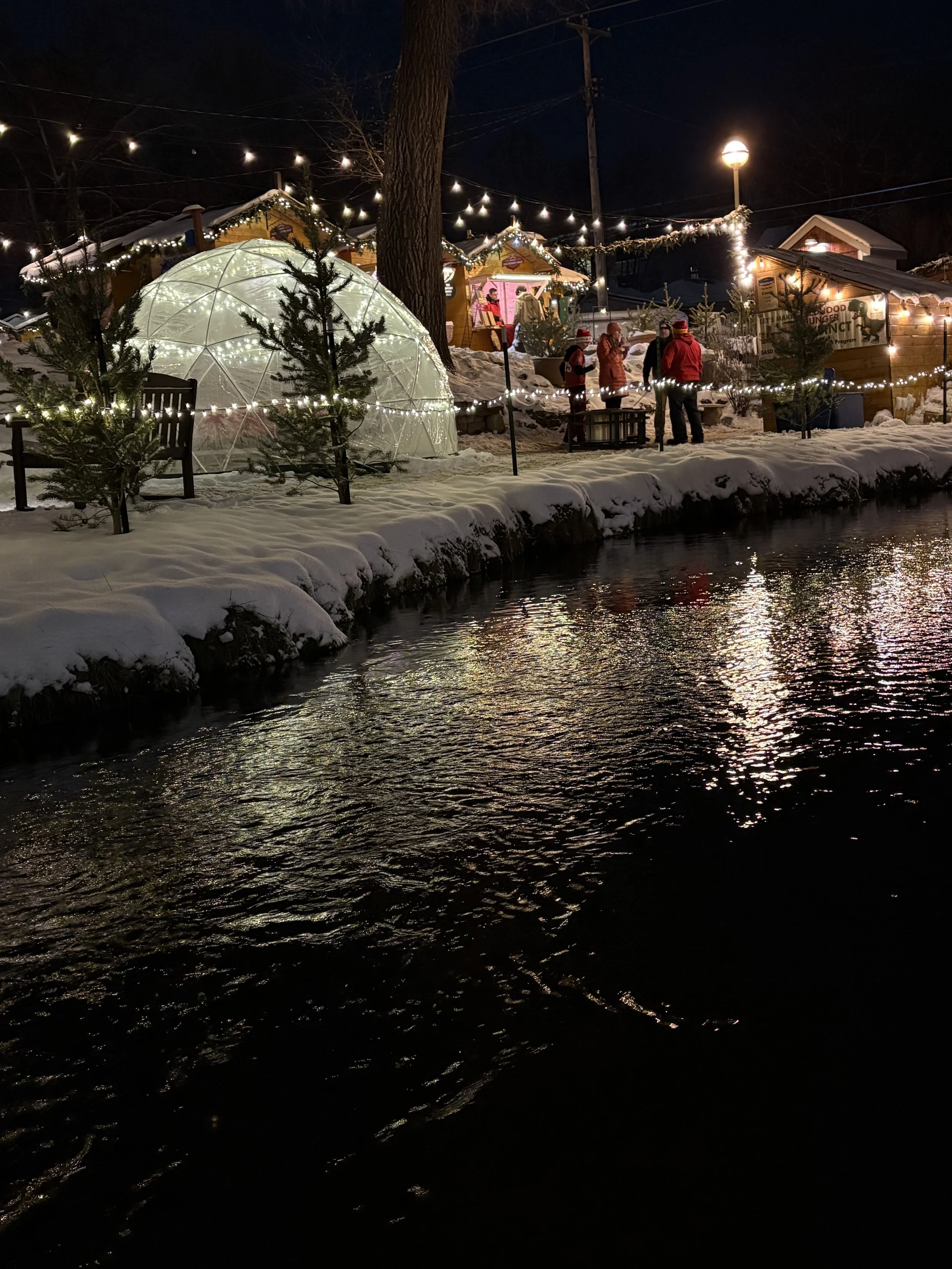 Night scene of a snow-covered outdoor area decorated with string lights. Small Christmas trees and a glowing geometric dome structure are visible. Several people dressed in winter clothing are gathered near small wooden cabins, with a body of water in the foreground reflecting the lights.