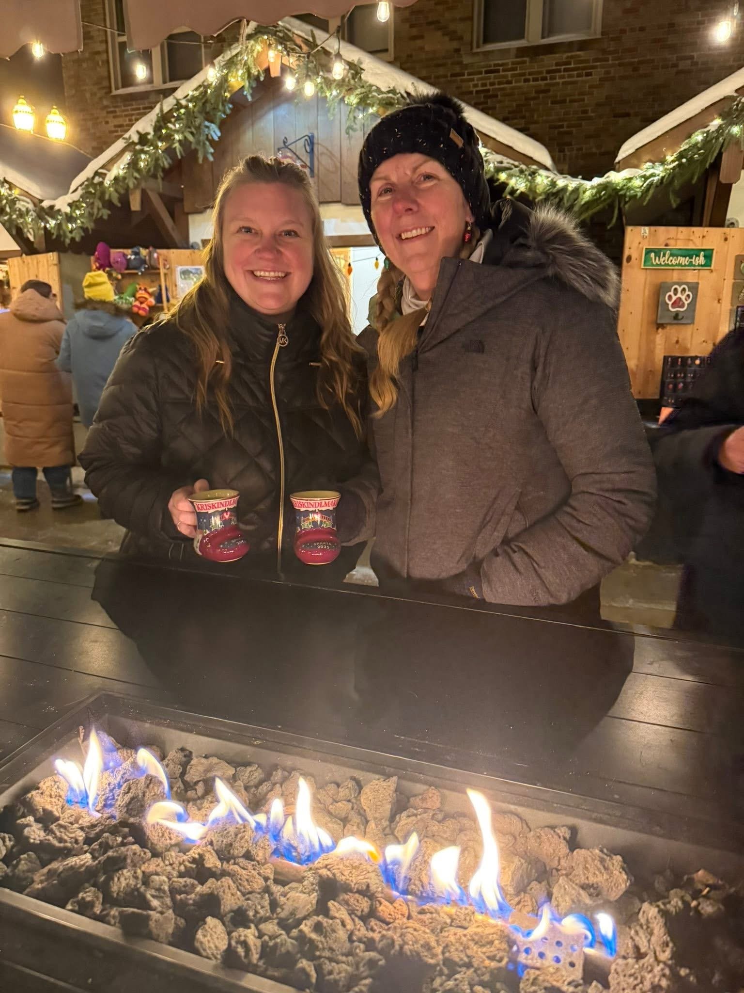 Two women smiling and holding cups at a festive outdoor setting with string lights and decorated wooden structures. In the foreground, a fire burns among rocks.