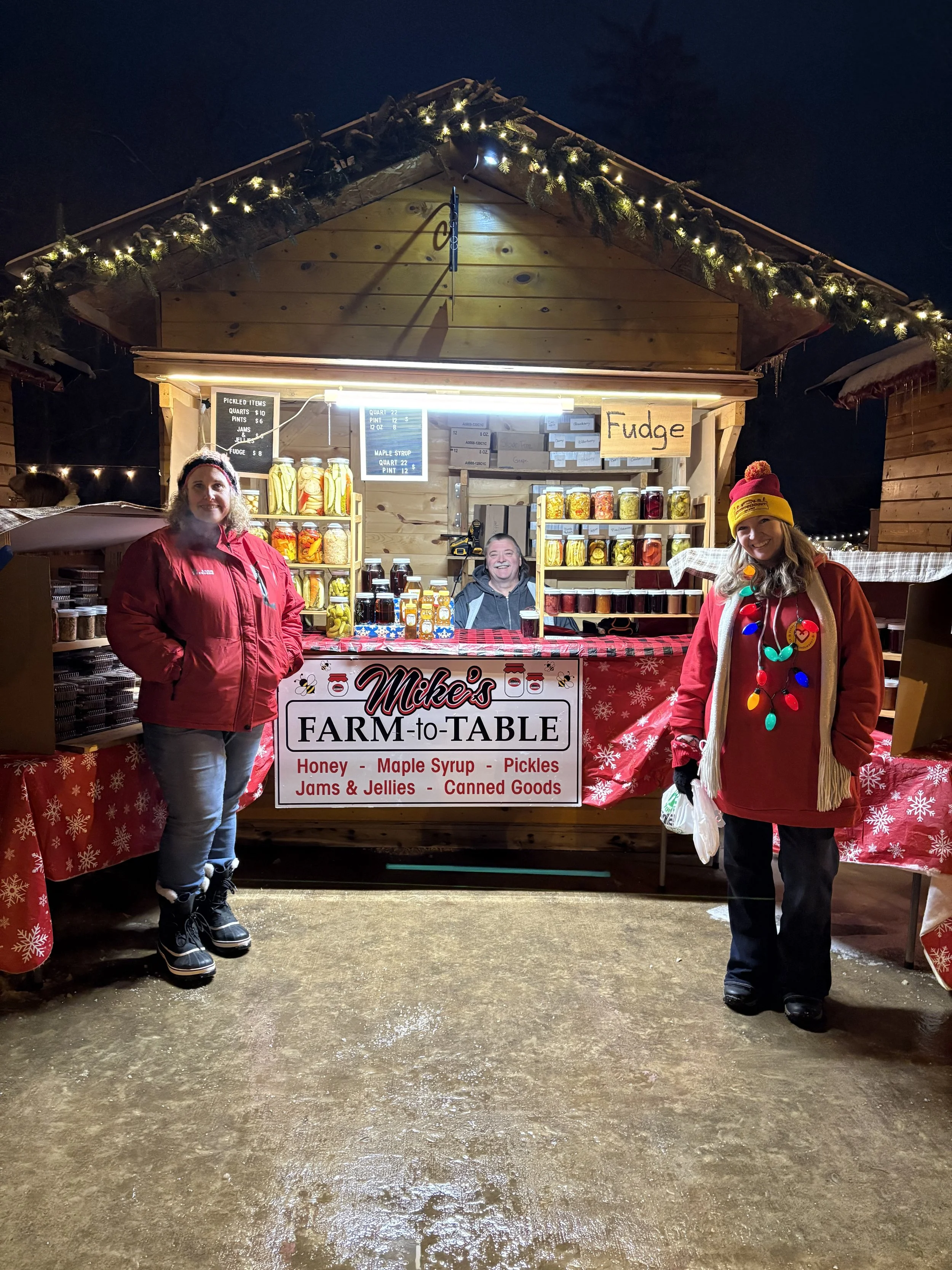 Two women in winter clothing standing in front of a wooden farm stand decorated with holiday lights and greenery, selling honey, maple syrup, pickles, jams, jellies, and canned goods at an outdoor market at night.