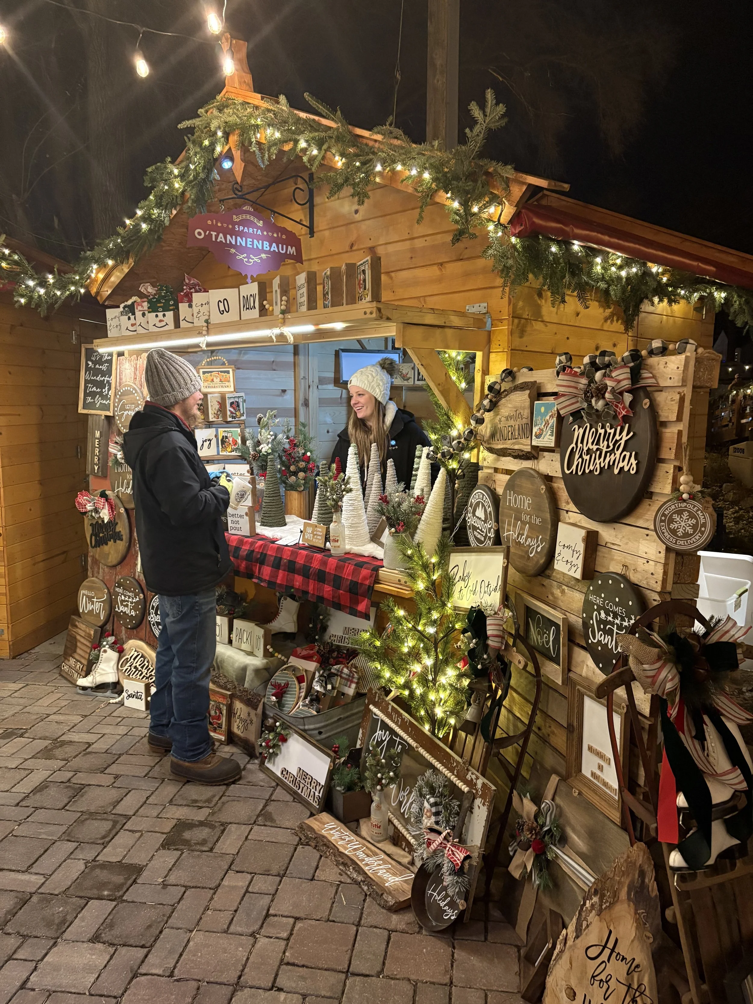 A Christmas market stall decorated with holiday signs, ornaments, and greenery. Two people are engaged in conversation, one man and one woman, both dressed warmly in winter clothing, at night.