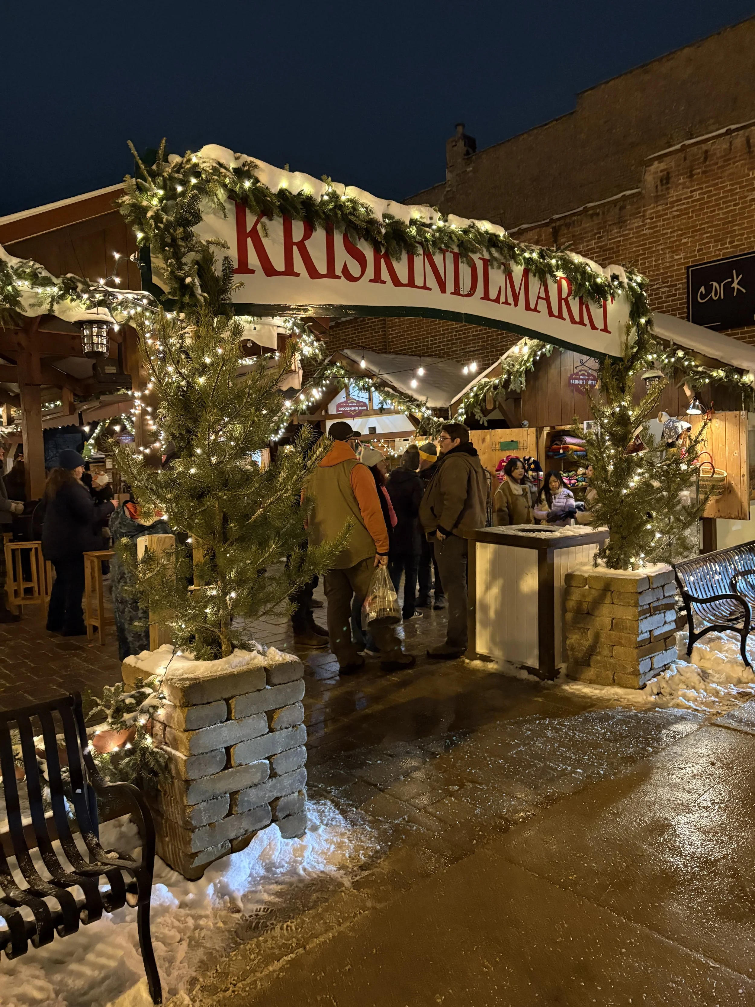 Nighttime scene at a Christmas market with people gathering under a festive sign that reads 'KRISKRINGLEMARKT'. The market is decorated with string lights and green garlands, with snow on the ground and on top of the market stall.