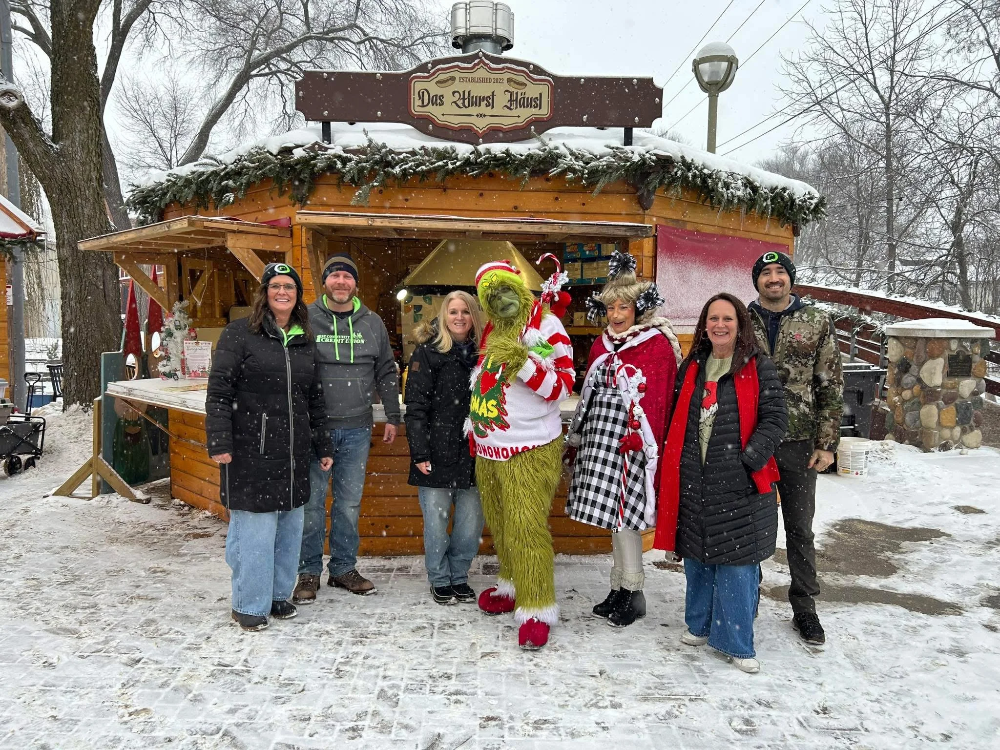 A group of six people dressed in winter clothing standing in front of a wooden food stand decorated with evergreen branches and a sign that reads 'Das Wurst Haus,' with snow falling.