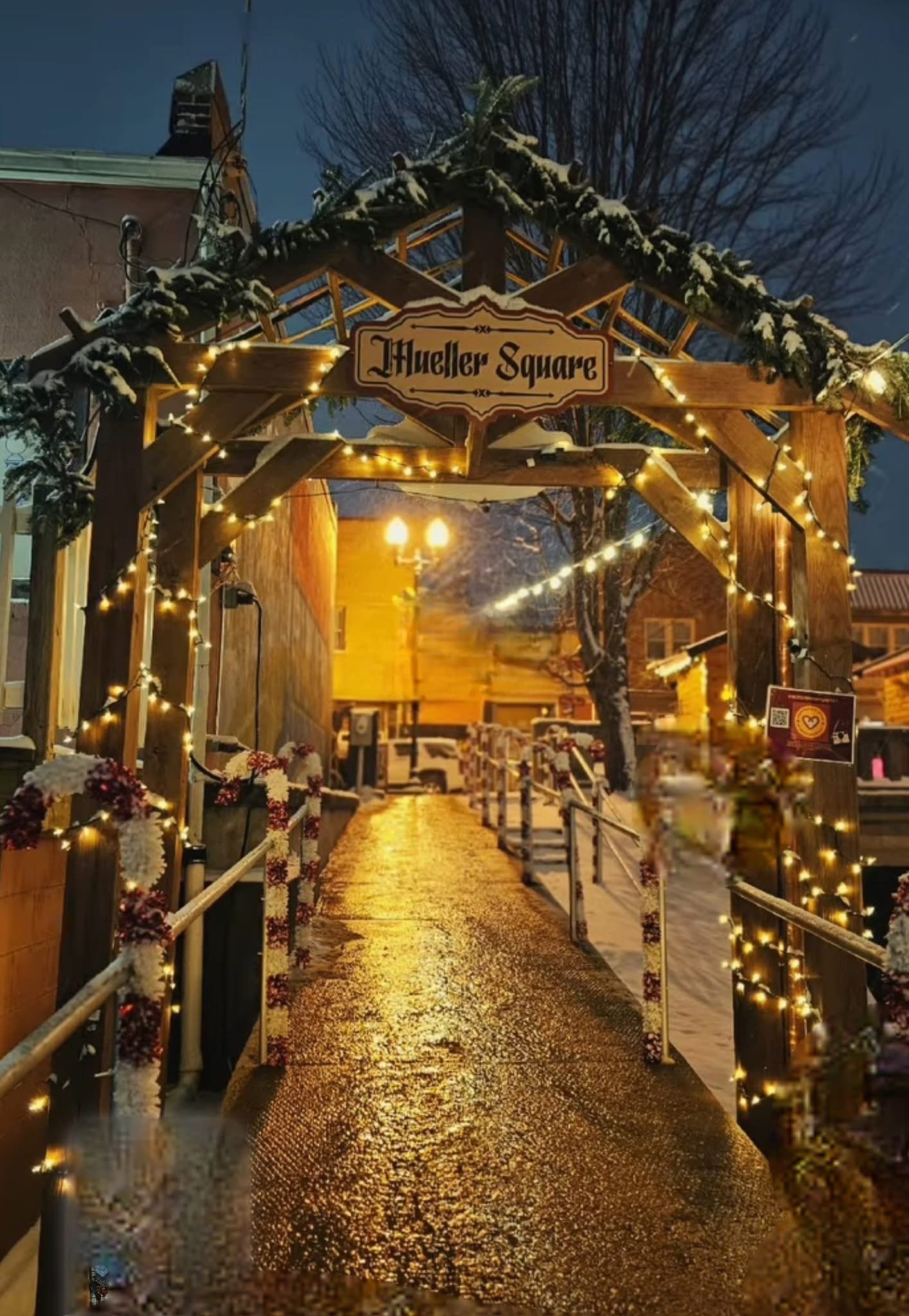 Night view of a street decorated with Christmas lights, snow, and a sign that reads 'Mueller Square.'