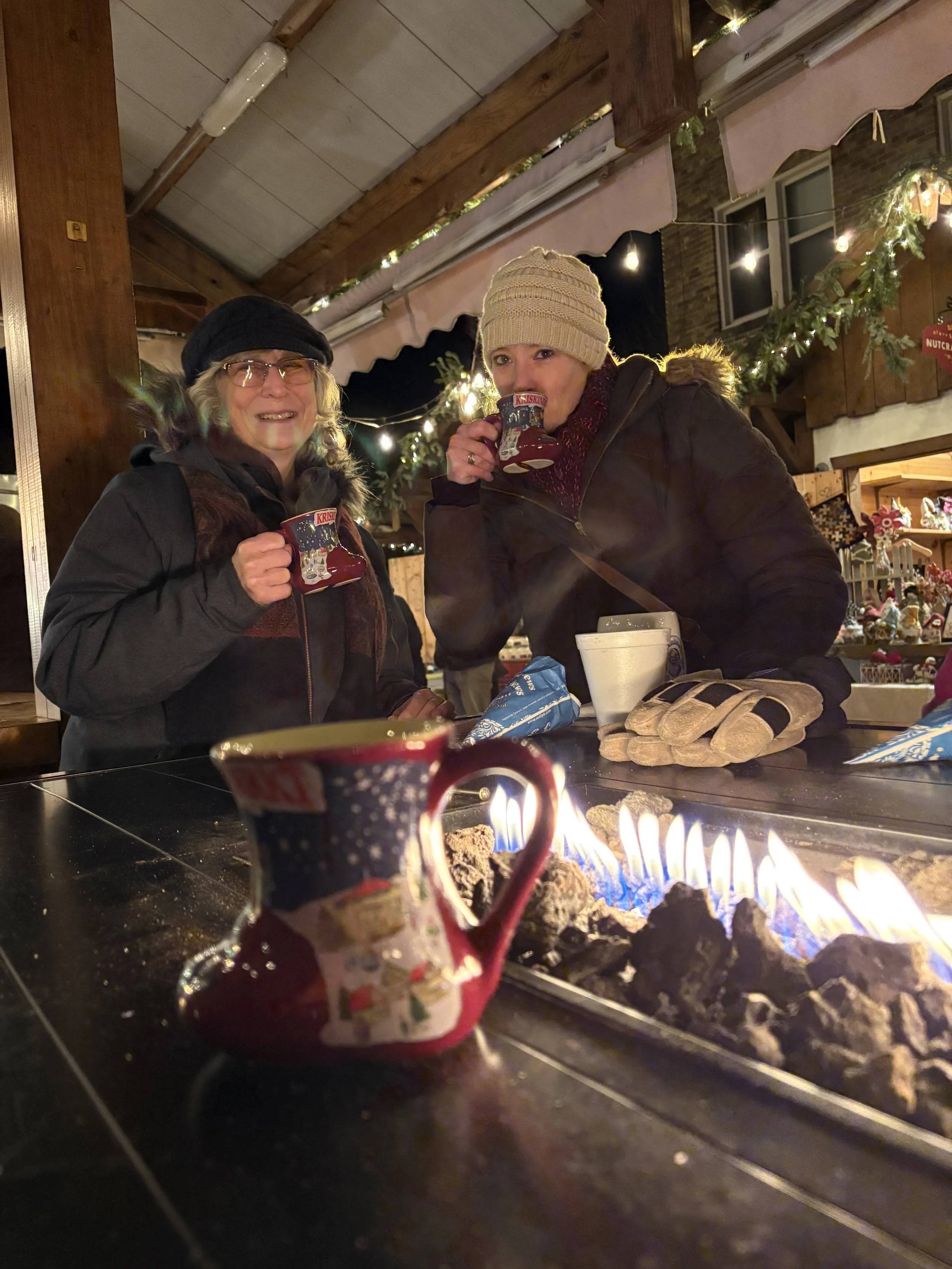 Two women dressed in winter clothing, including hats and gloves, sitting at a table with Christmas mugs, in a festive decorated setting with string lights and greenery, near a fire pit.