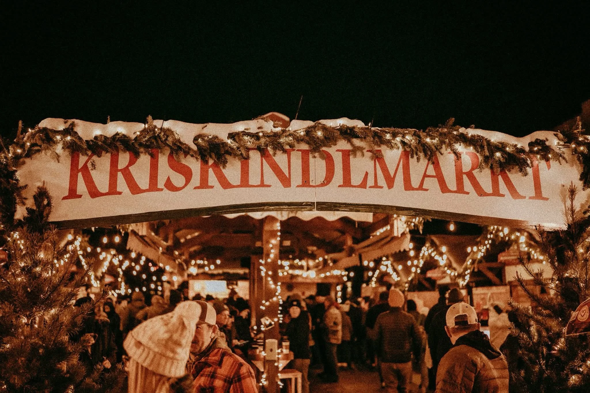 A crowded Christmas market at night with festive lighting, decorated with greenery and a large sign reading 'KrisKindlMart' in red letters.