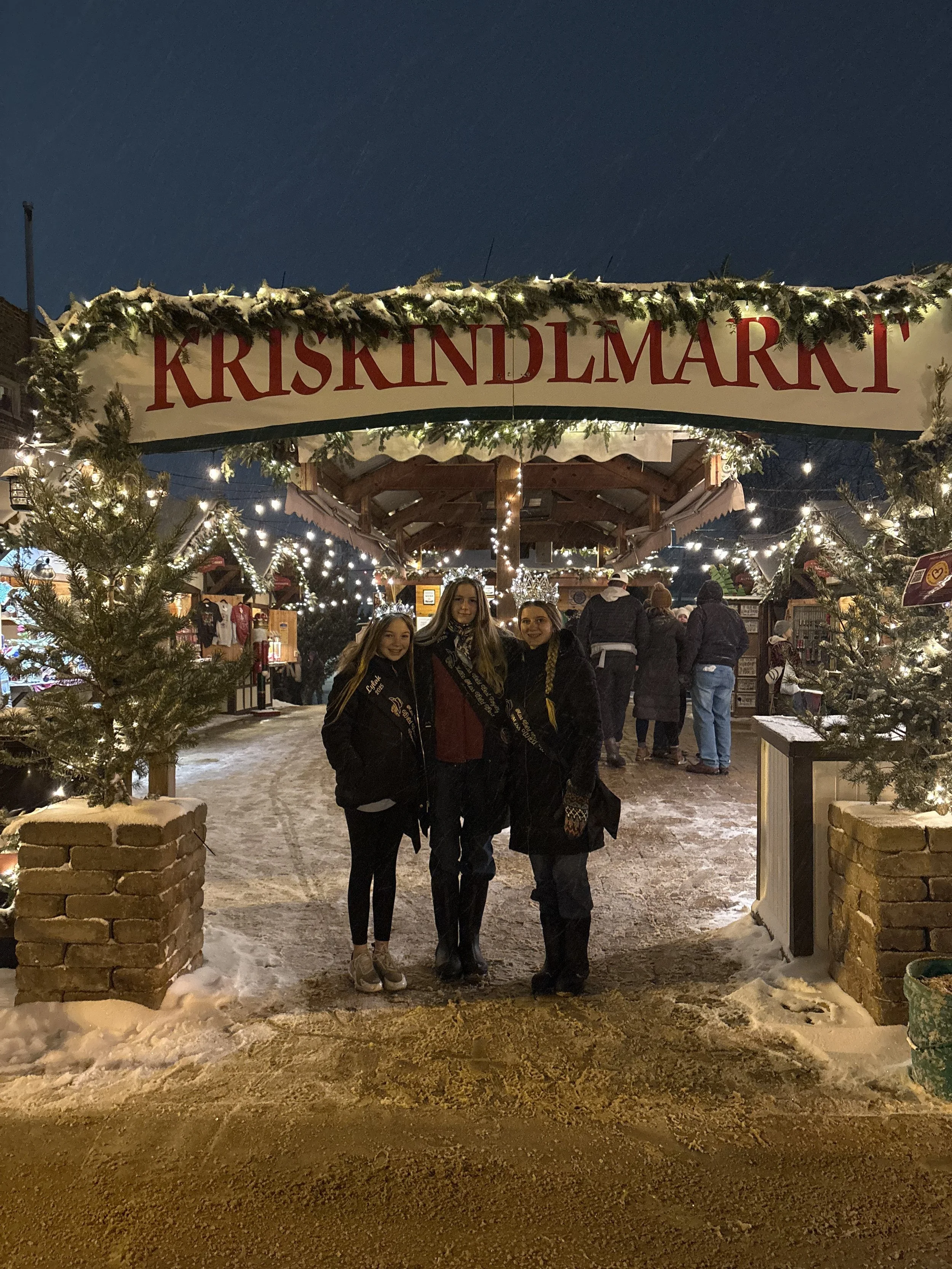 Three women standing in front of a Christmas market entrance decorated with lights and greenery, with snow on the ground at night.