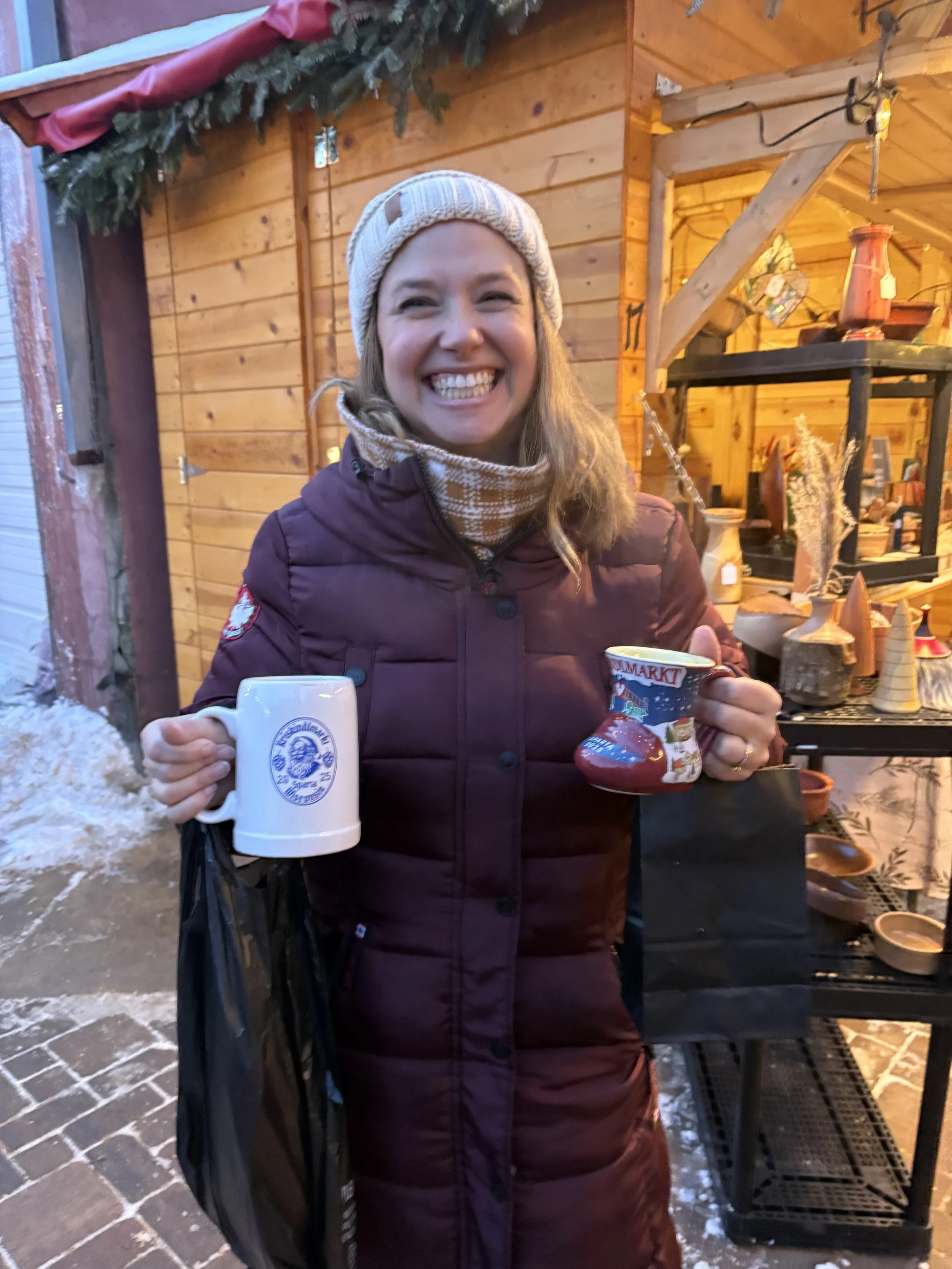 Smiling woman in winter clothing holding a coffee mug and a small Christmas-themed sock with a snowman design, standing outdoors near a decorated wooden booth with holiday decorations.