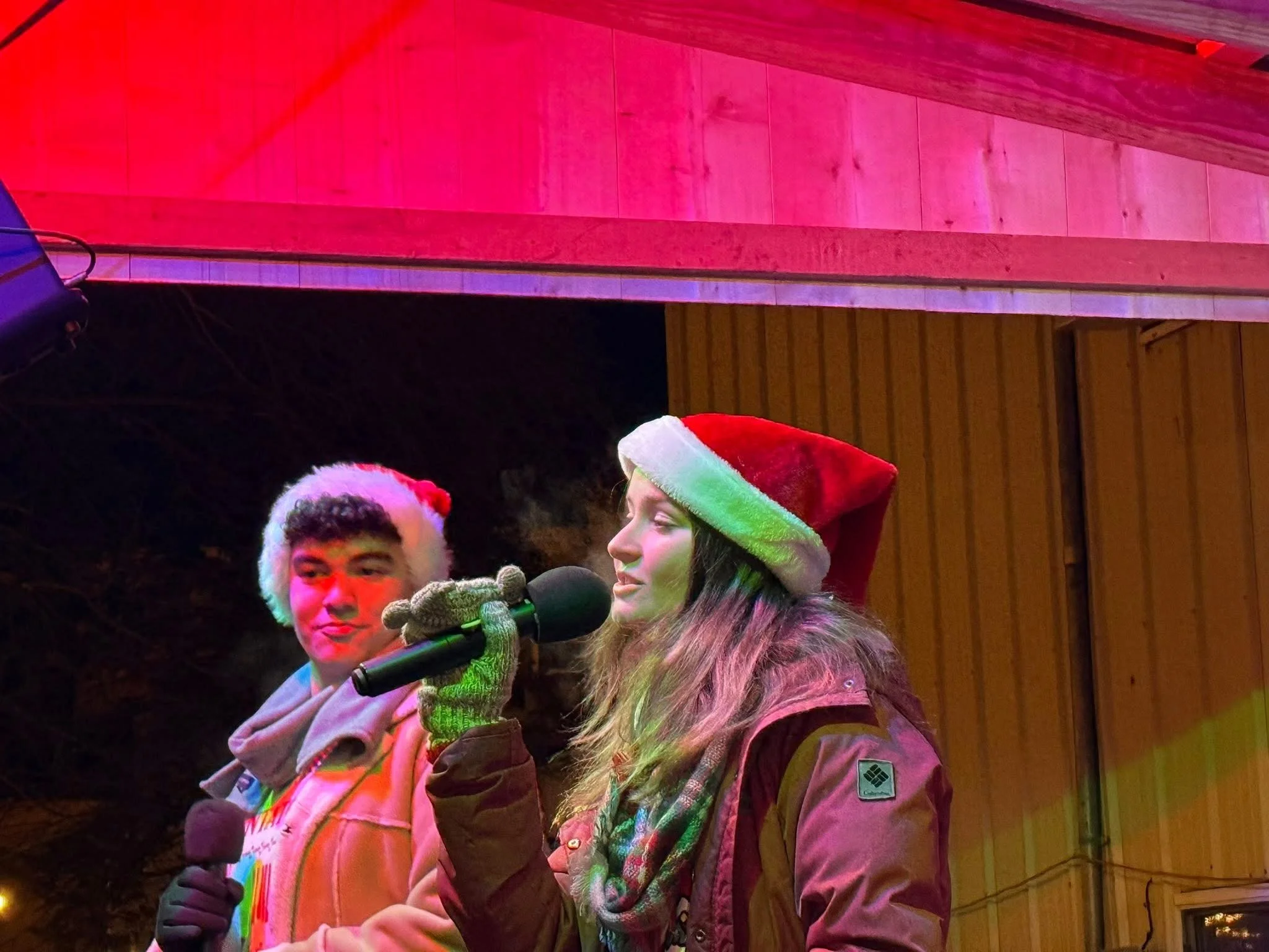 A group of people, possibly at a holiday or Christmas event, with one person in a Christmas-themed hat speaking into a microphone. The scene appears to be indoors with a wooden wall backdrop, and there are multi-colored lights creating a festive atmosphere.