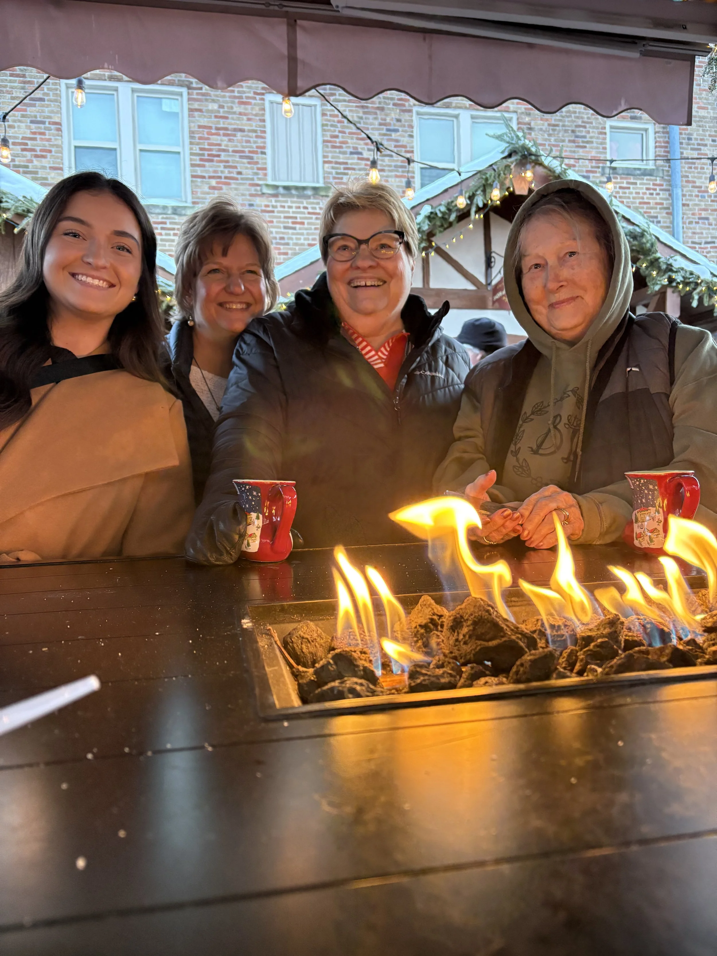 Four women sitting outdoors at a table with a fire pit in front of them, smiling, under string lights with a brick building in the background.