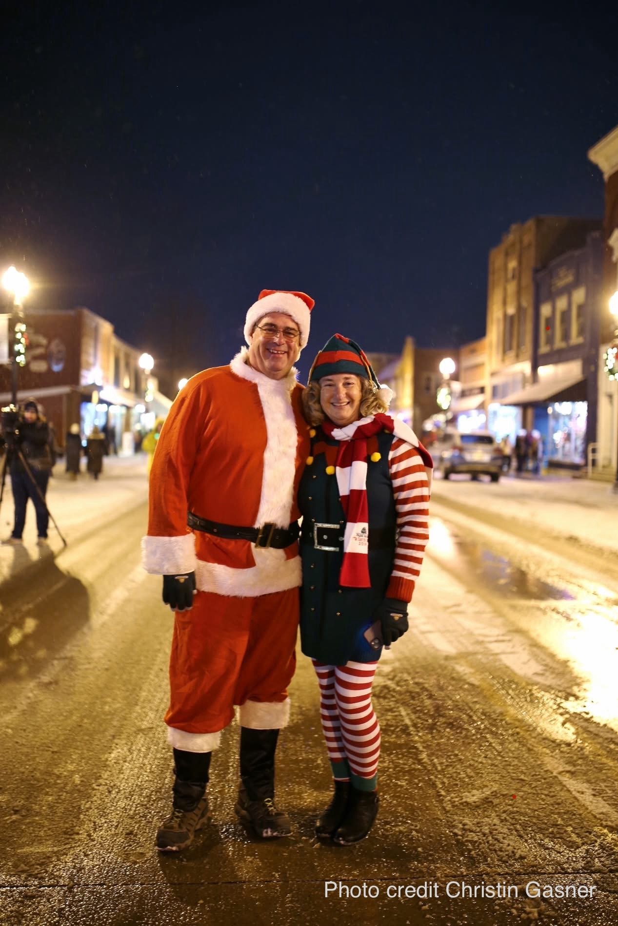 Man dressed as Santa Claus and woman dressed as an elf standing together on a decorated street at night, smiling for the camera.