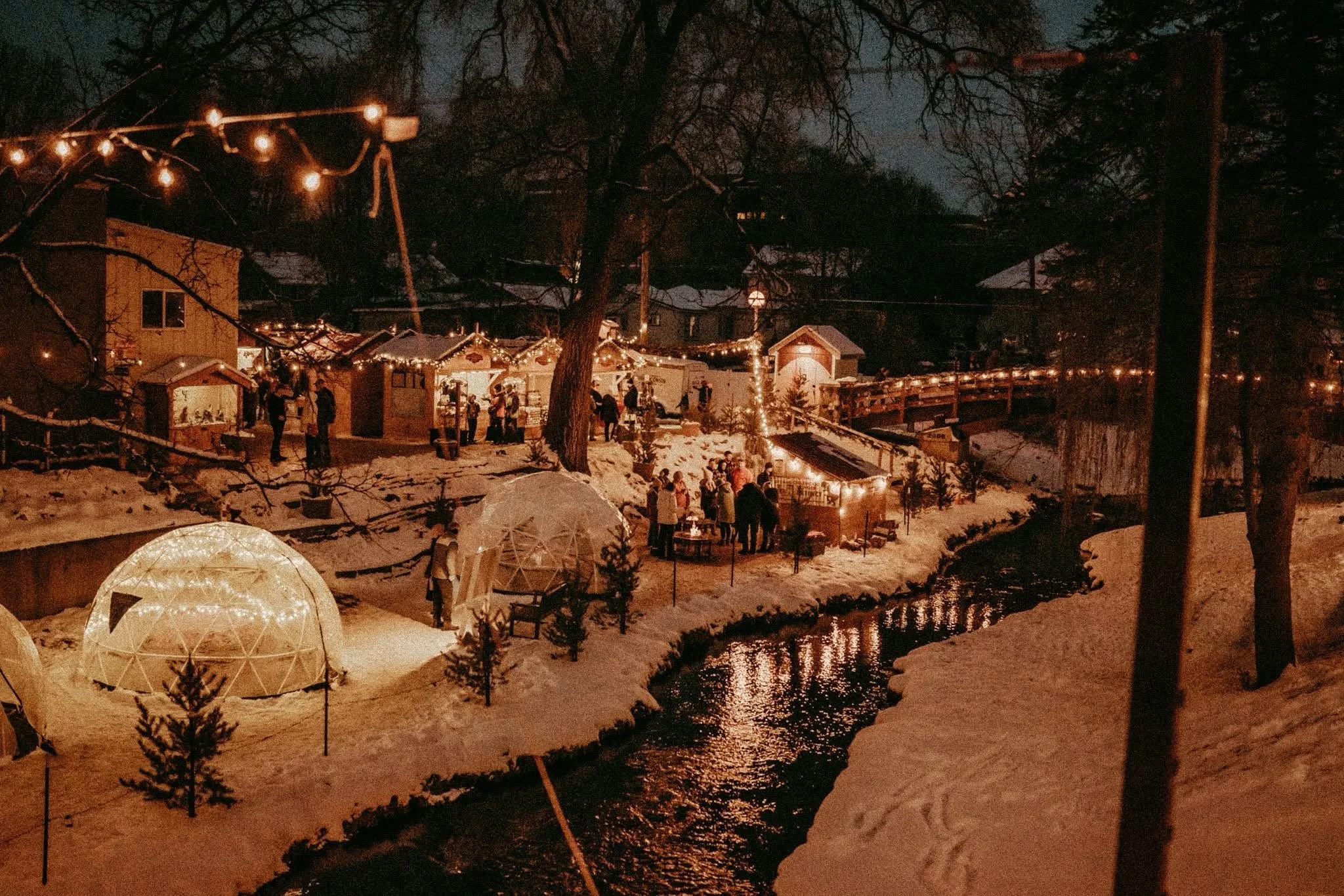 Night scene of an outdoor winter celebration with string lights, small houses or stalls decorated with lights, and people gathered around. Snow covers the ground near a river or creek, reflecting the warm glow of lights. Snowy trees frame the scene.