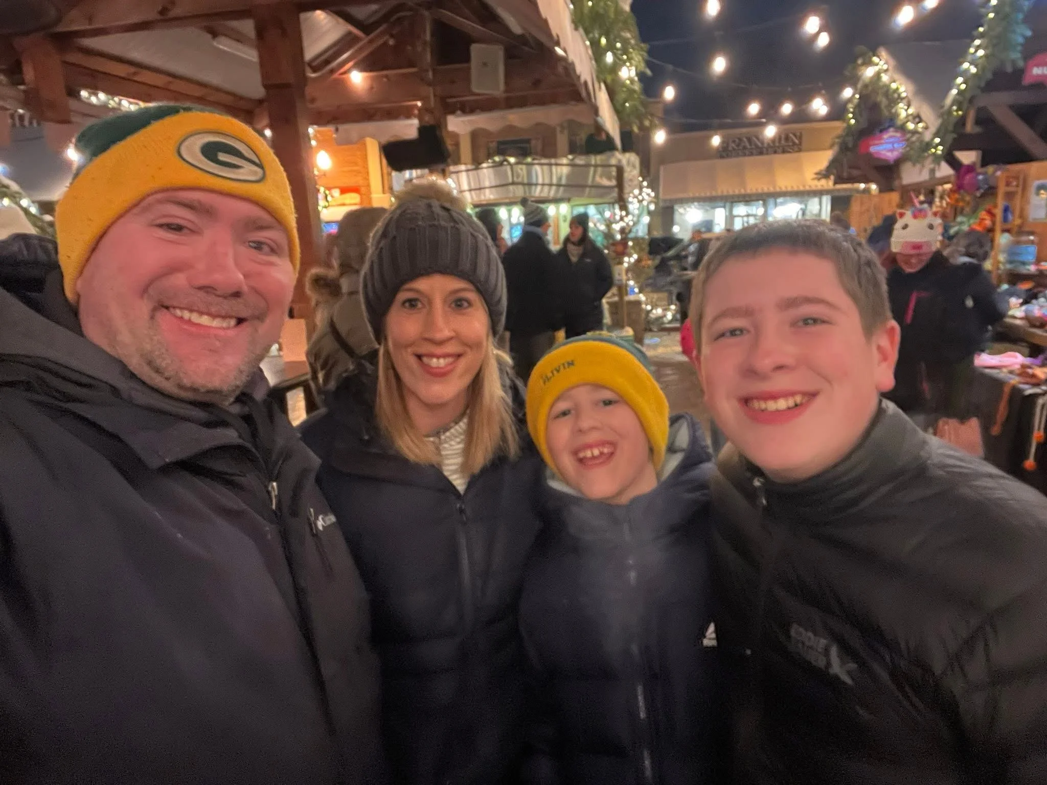 A group of four smiling people taking a selfie at a festive outdoor market decorated with string lights and holiday ornaments during the evening.