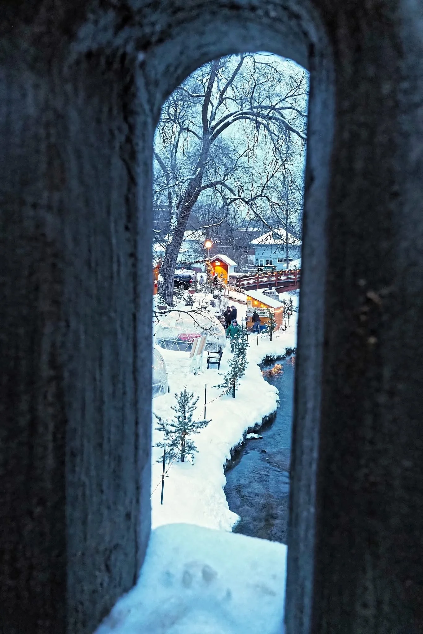 View through a small, arched opening in a wall showing a snowy landscape with a tree, houses, and a small creek at dusk.