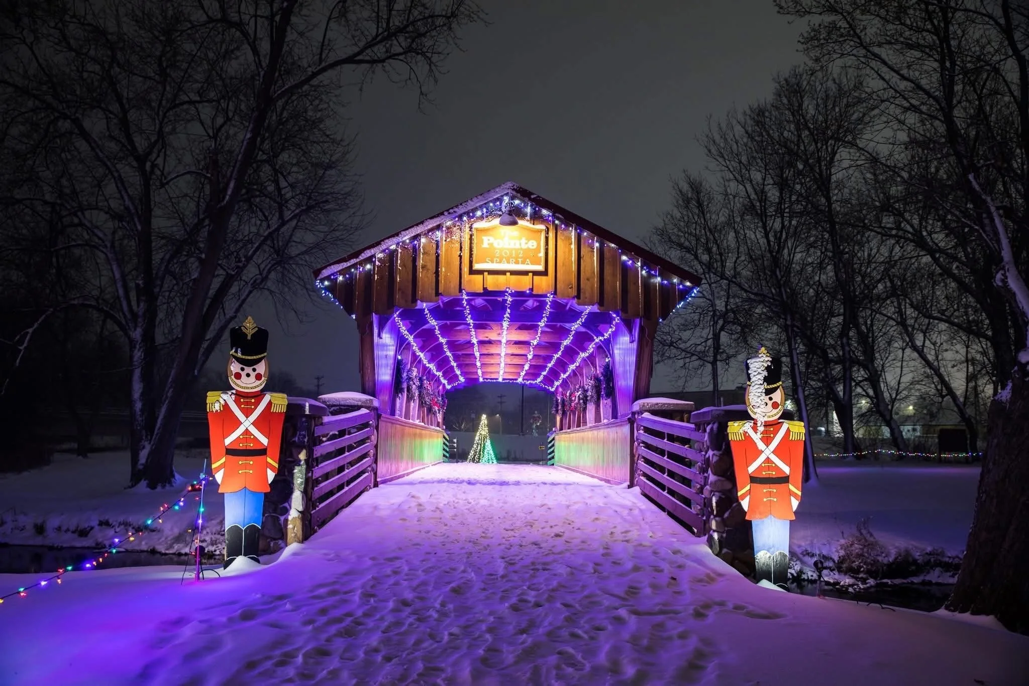 A wooden covered bridge decorated with purple and white Christmas lights, with toy soldier figures at each entrance, on a snowy night.