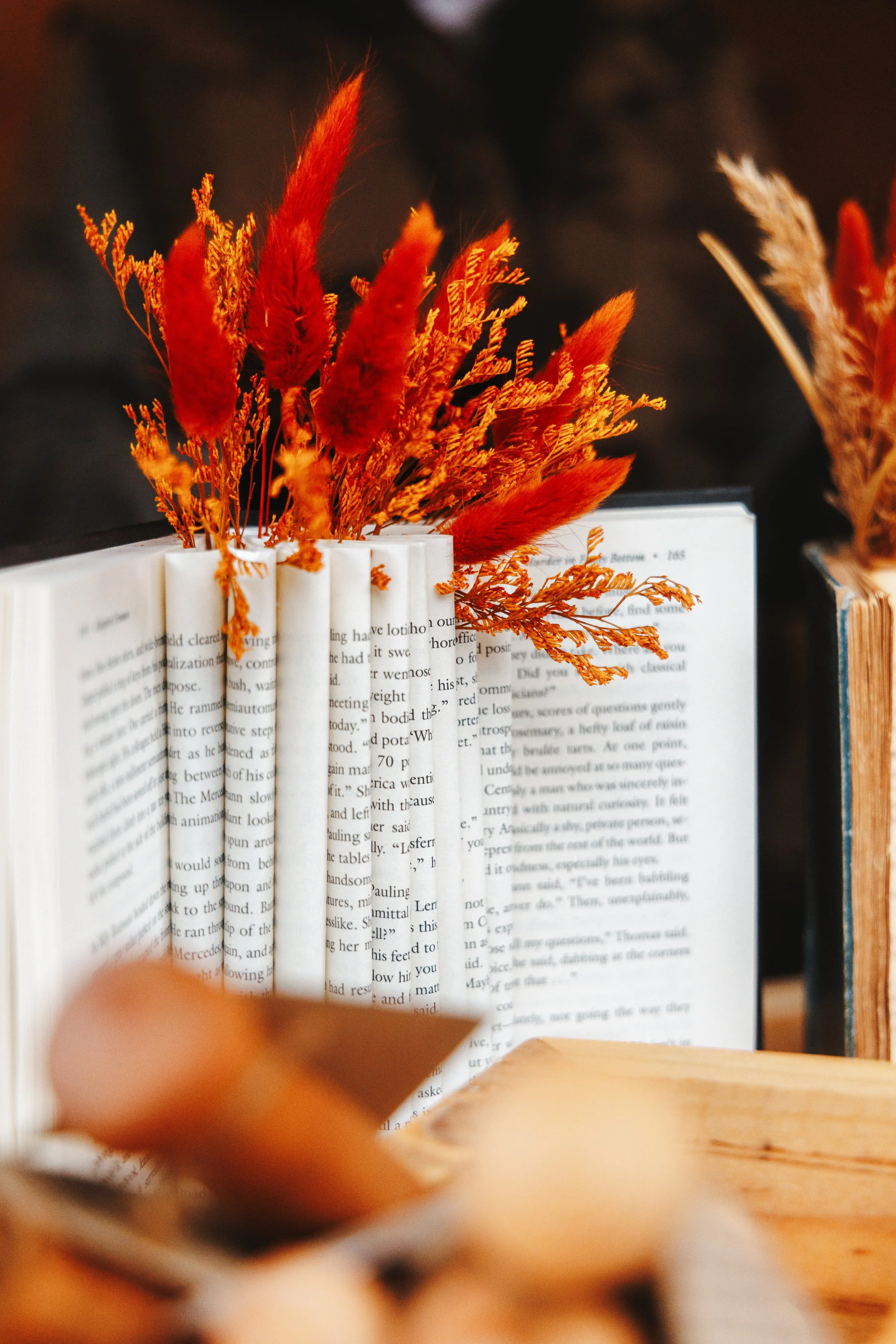 A close-up of a book with orange and red dried flowers placed inside it, seen through the pages. The background is blurred, with warm lighting highlighting the flowers.