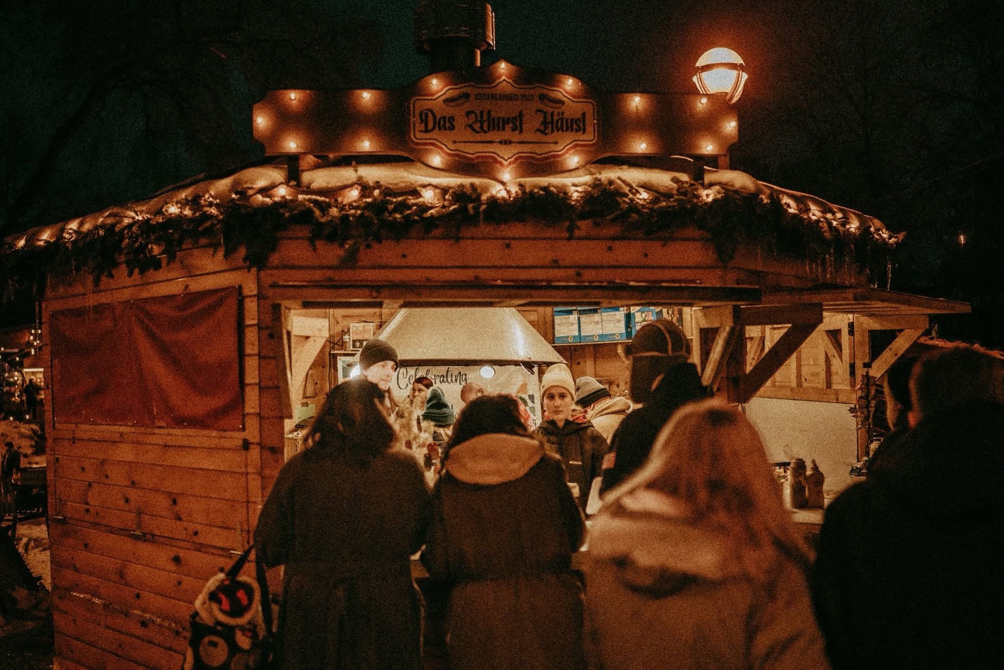 Nighttime scene at a festive outdoor market with a wooden food stand decorated with garlands, illuminated by warm string lights and a lamp, with people gathered around and a sign reading "Das Urst Häusl."