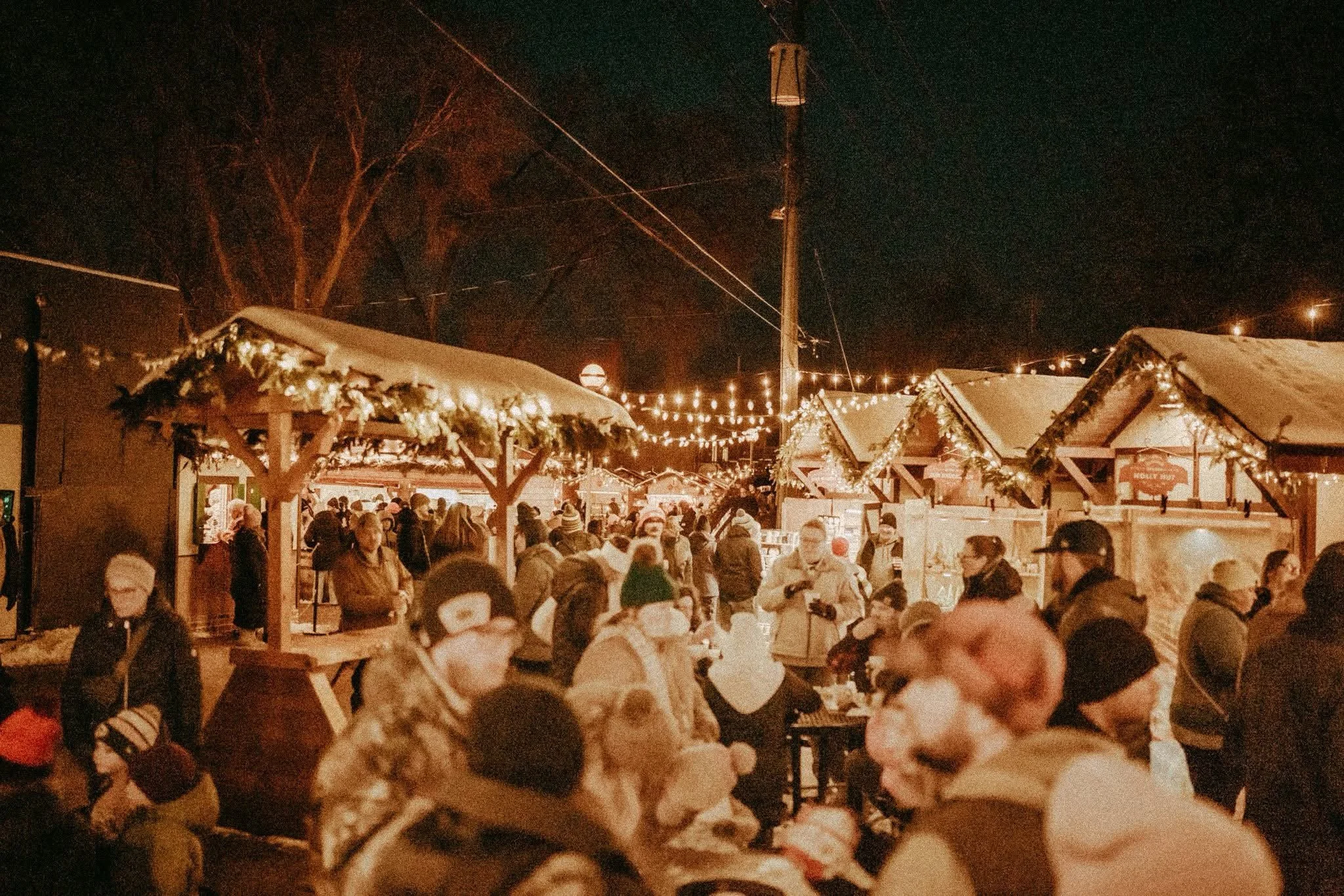 A crowded outdoor Christmas market at night with string lights, snow-covered rooftops, and many people shopping and walking around.