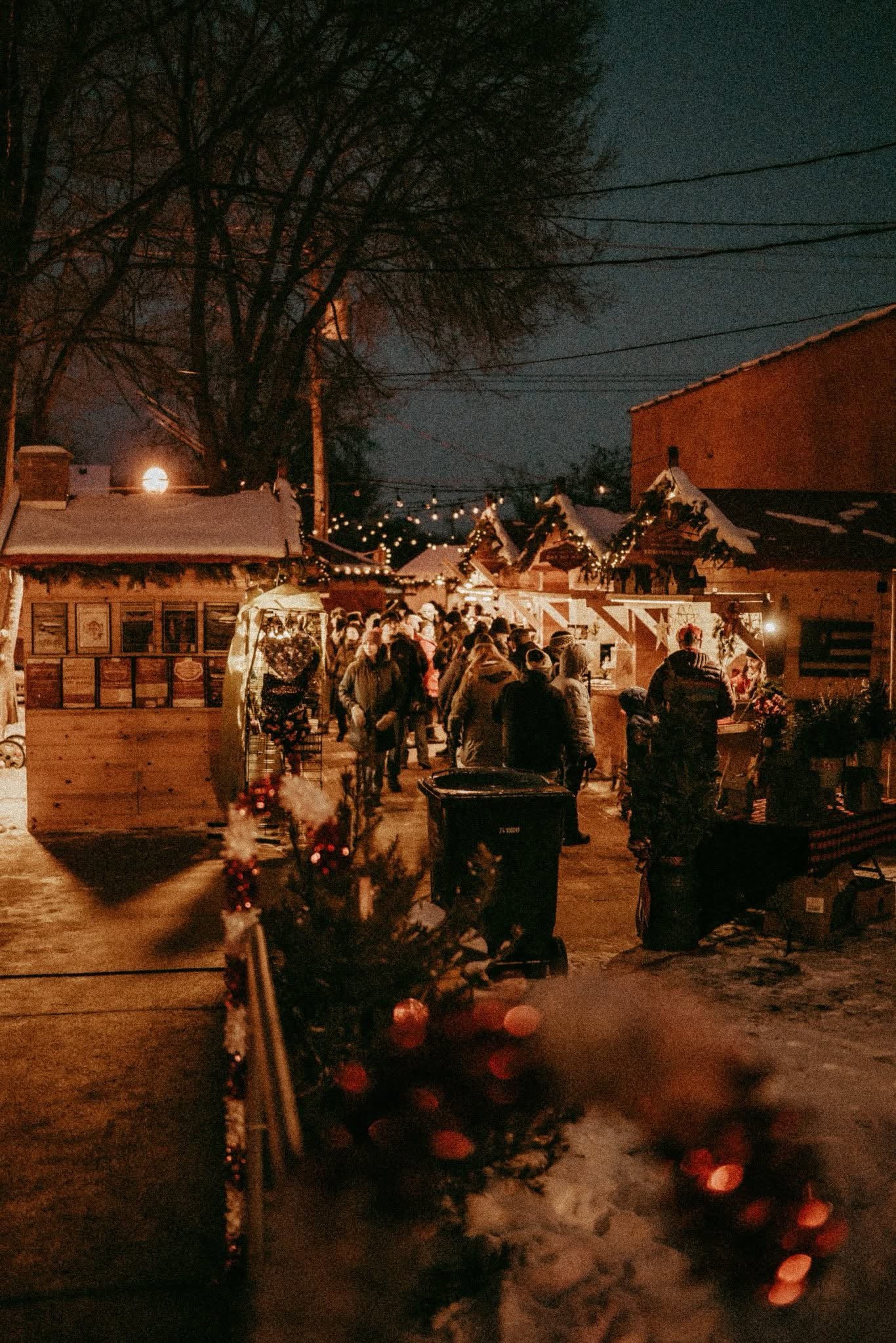 A bustling outdoor holiday market at night with warm string lights, wooden stalls, and a crowd of people dressed in winter clothing. Snow covers the ground and roofs, with a large tree and a full moon visible in the dark sky.