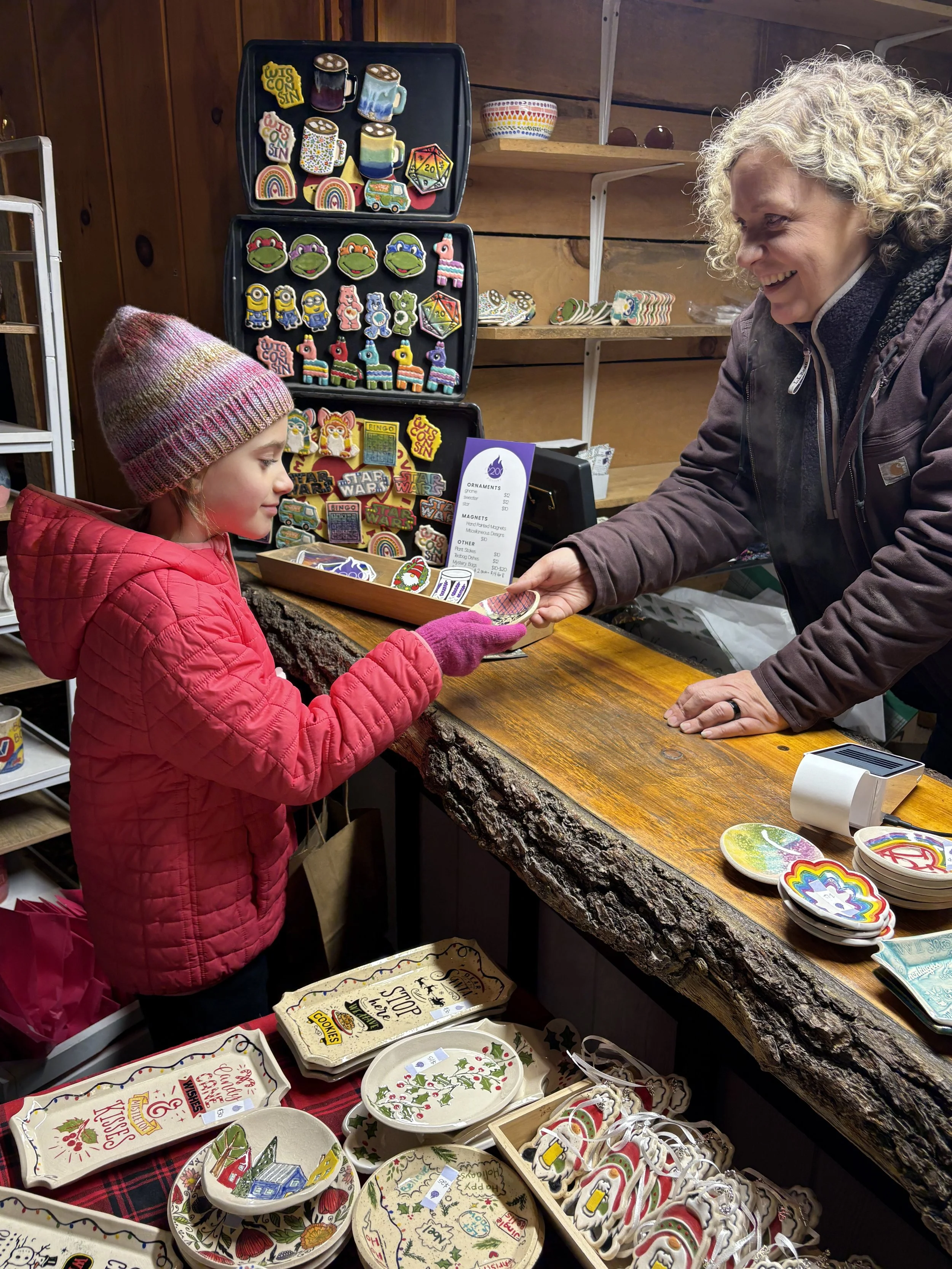 A young girl in a pink jacket and knit hat buying holiday-themed ceramic and decorative items from an older woman at a craft booth. The booth displays various colorful holiday-themed items, including plates, ornaments, and patches with designs like rainbows, animals, and Santa Claus.