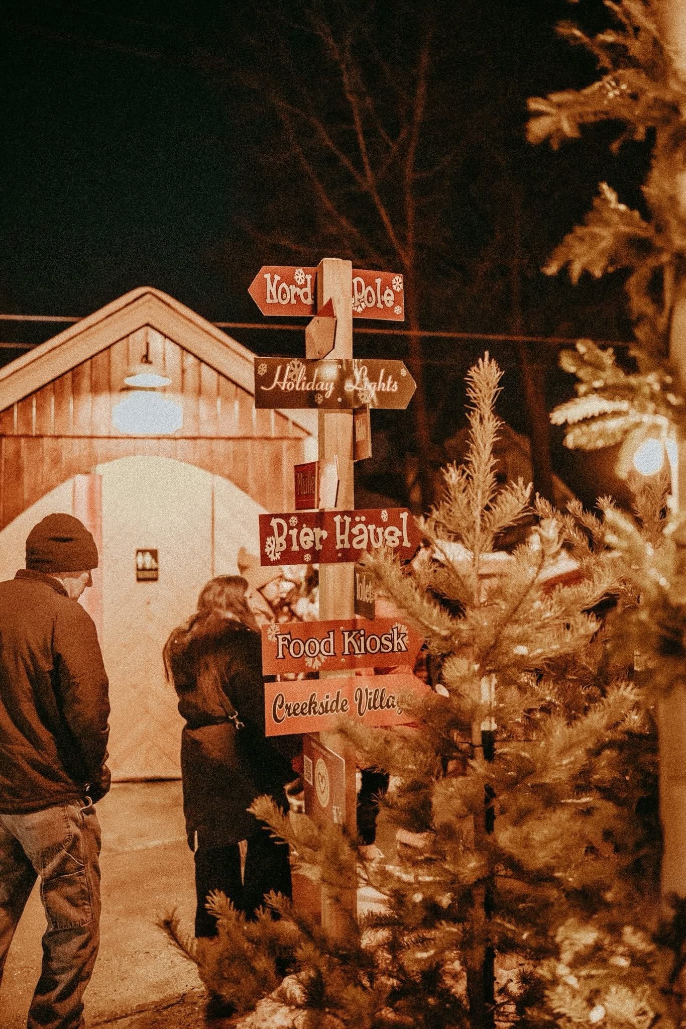 Night scene with Christmas tree and a signpost showing directions to various holiday attractions, including Nord Pole, Holiday Lights, Bier Häusel, Food Kiosk, and Creekside Villa, with a building, trees, and people's silhouette in the background.