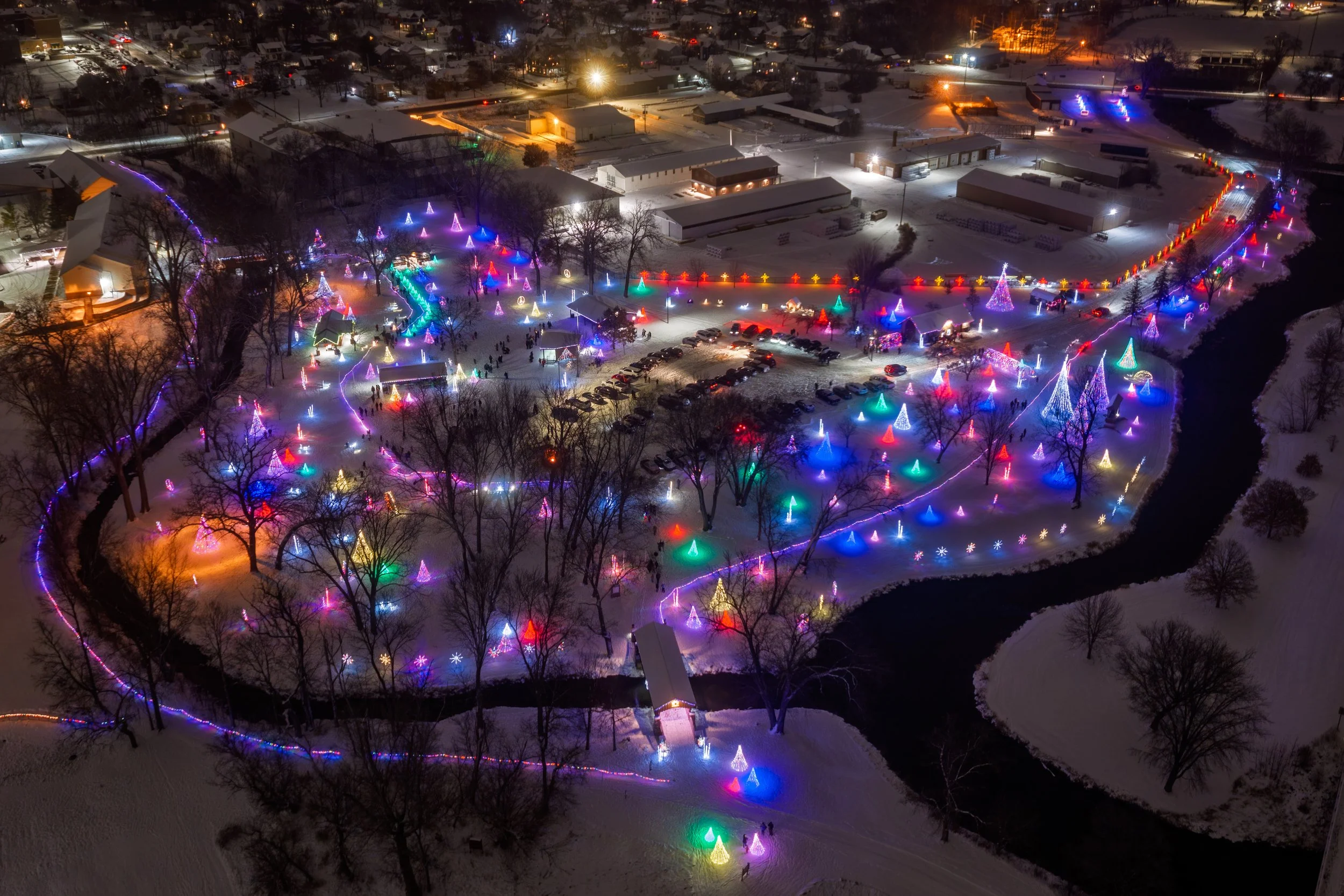Night aerial view of a town with snow-covered ground, showcasing colorful Christmas lights and decorations, including lit Christmas trees and strings of lights along pathways, with a parking lot and buildings in the background.
