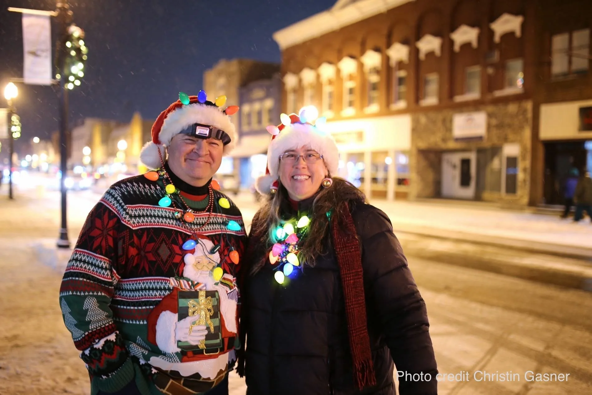 A man and a woman are standing outside on a snowy street at night, smiling and dressed festively for Christmas. The man is wearing a Santa hat decorated with Christmas lights, a black and red holiday sweater, and has garland around his neck. The woman is wearing glasses, a Santa hat decorated with Christmas lights, and holiday earrings, with a string of Christmas lights around her neck. There are buildings and streetlights in the background.