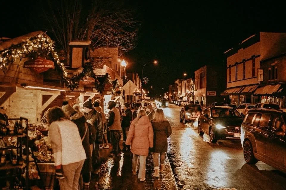 Night scene of a busy town street decorated for Christmas with holiday lights and garlands, showing people browsing outdoor market stalls and cars parked along the road.