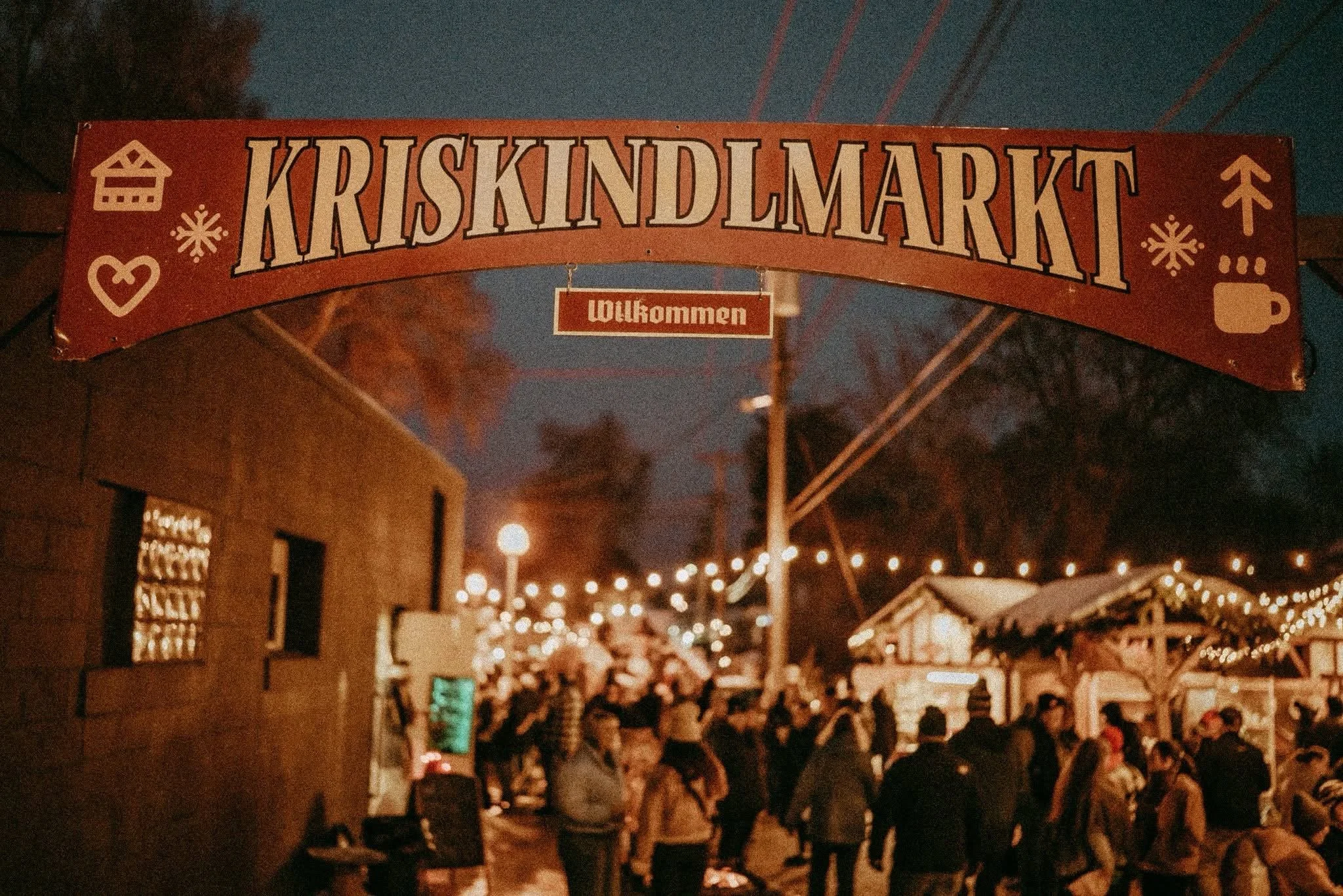 Nighttime scene of a crowd at a Christmas market with string lights, a large red sign reading 'Kriskindlmarkt' with decorative icons, and smaller sign saying 'Willkommen' underneath.
