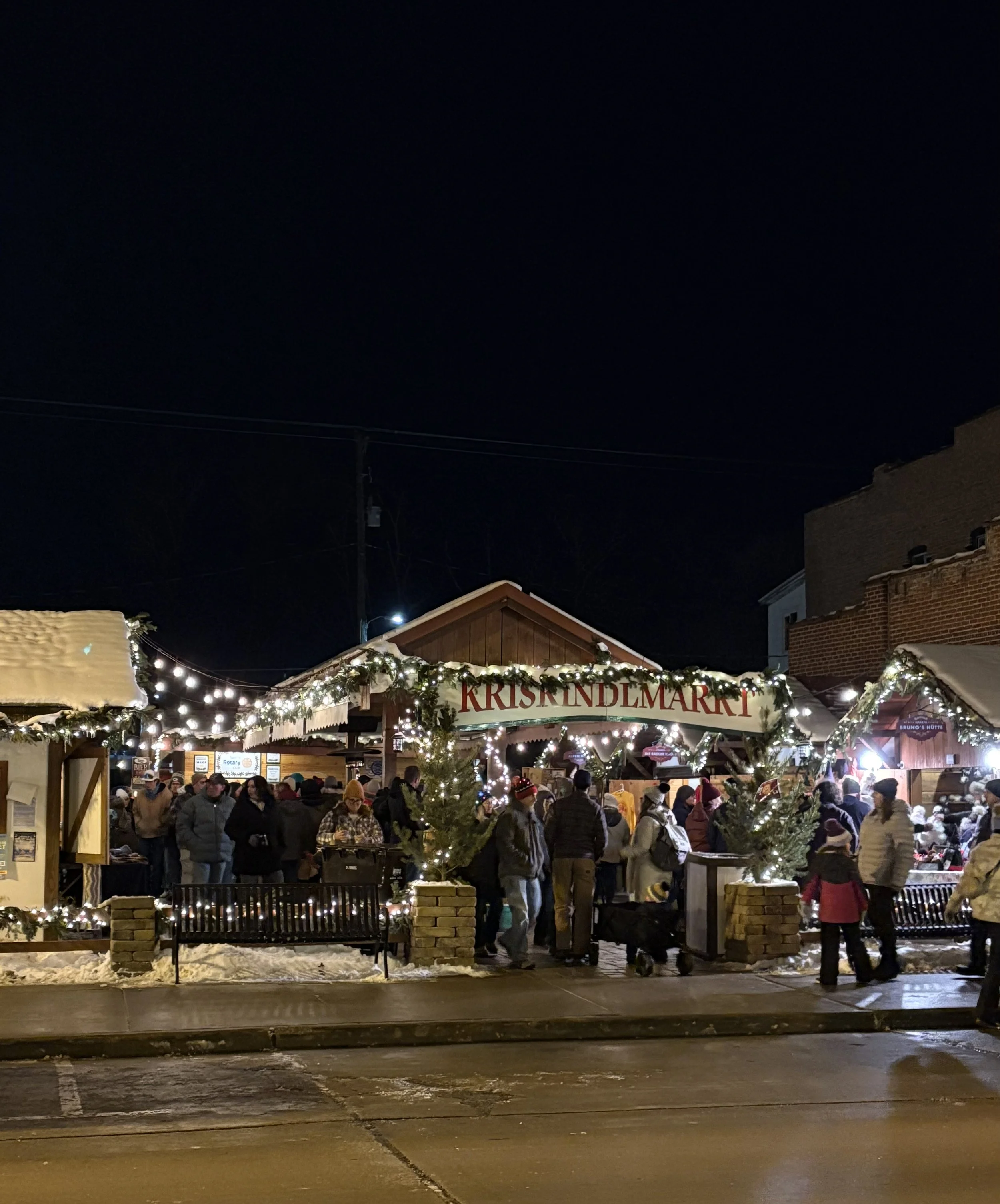 Nighttime scene at a Christmas market with people shopping under decorated stalls with string lights and a sign that reads 'KRISENDELMARKT'; snow on the ground and buildings.