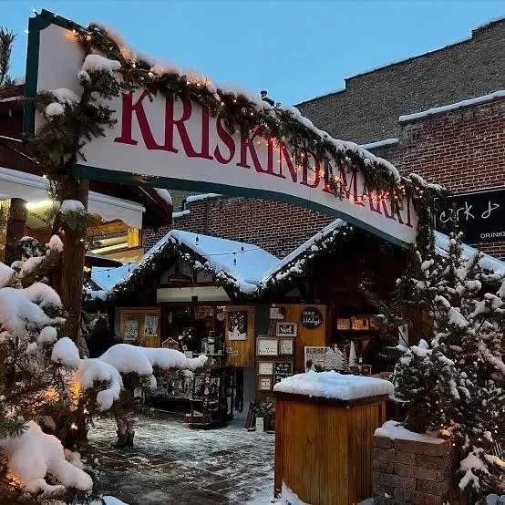 Snow-covered Christmas market entrance with a sign reading 'Kriskindlmarkt' and decorated evergreen branches hanging overhead.
