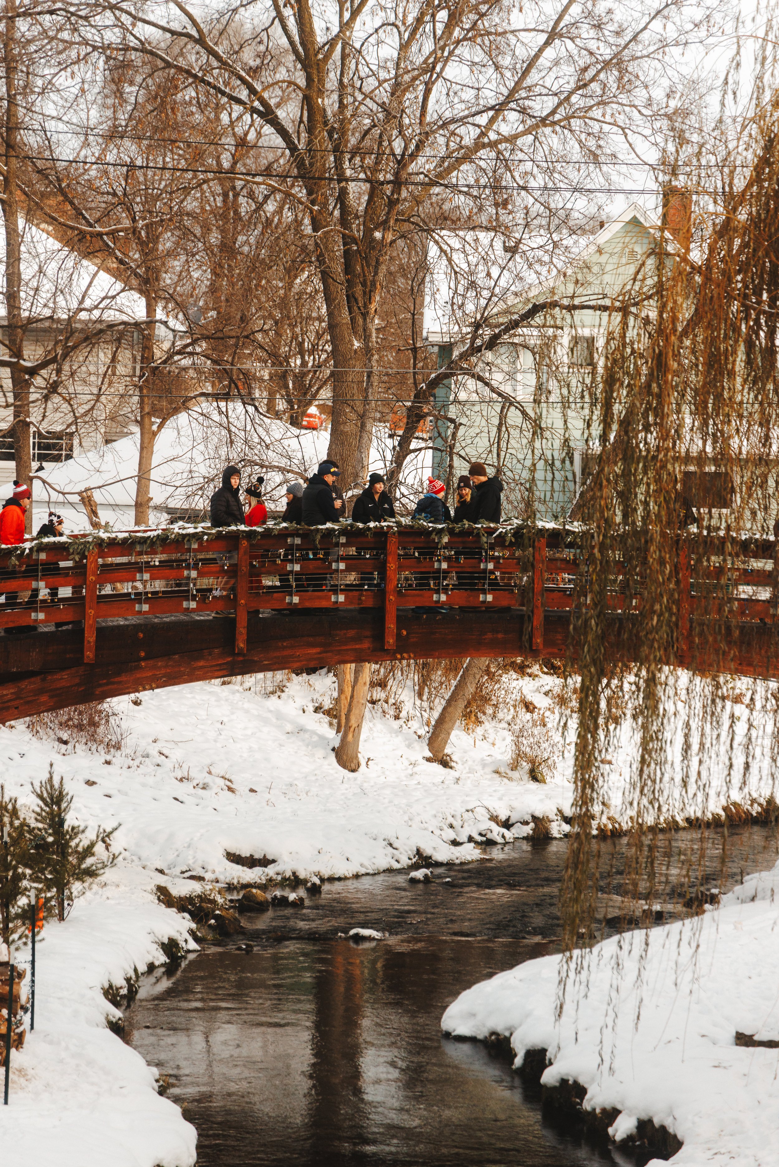 People gathered on a wooden bridge over a snow-covered creek in a winter landscape with leafless trees and houses in the background.