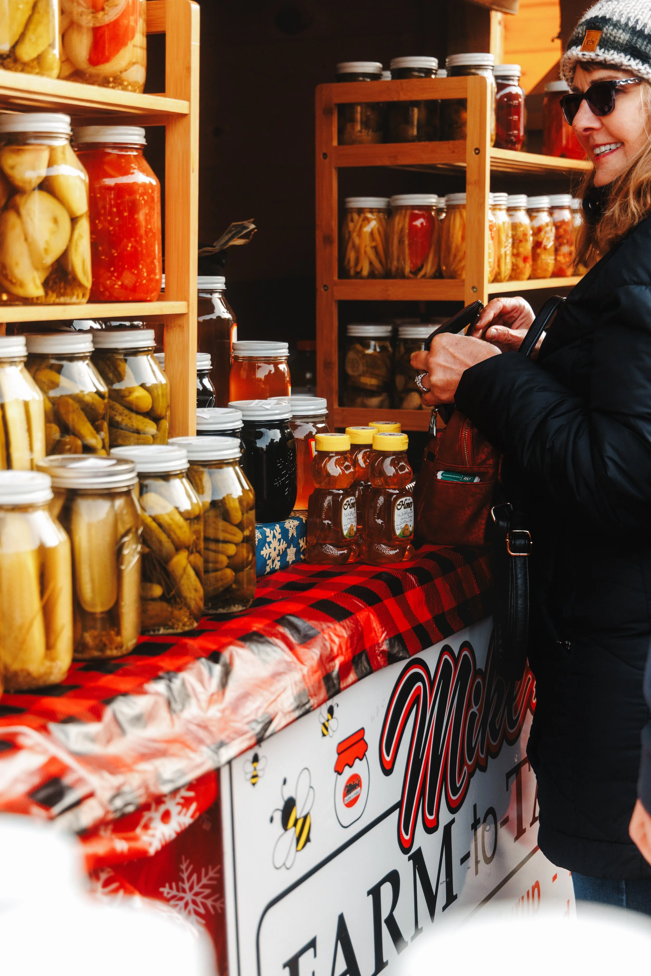A woman wearing sunglasses, a knit hat, and a black jacket shopping at a market stall with jars of pickled vegetables, jams, and honey on a red and black checkered tablecloth.