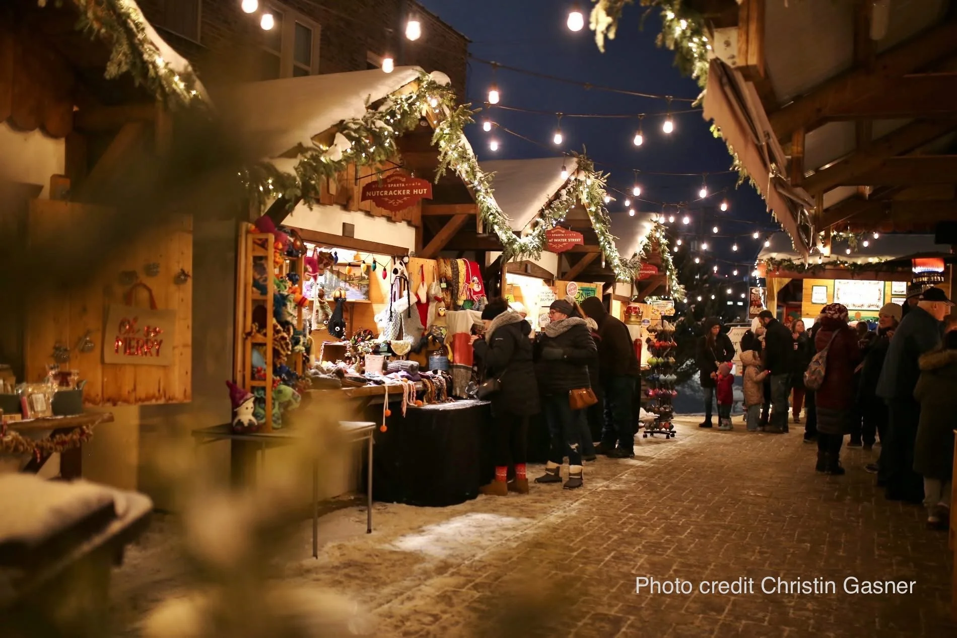Night scene at a Christmas market with decorated stalls, string lights, and holiday decorations. People are shopping and socializing, with snow on the ground and festive atmosphere.
