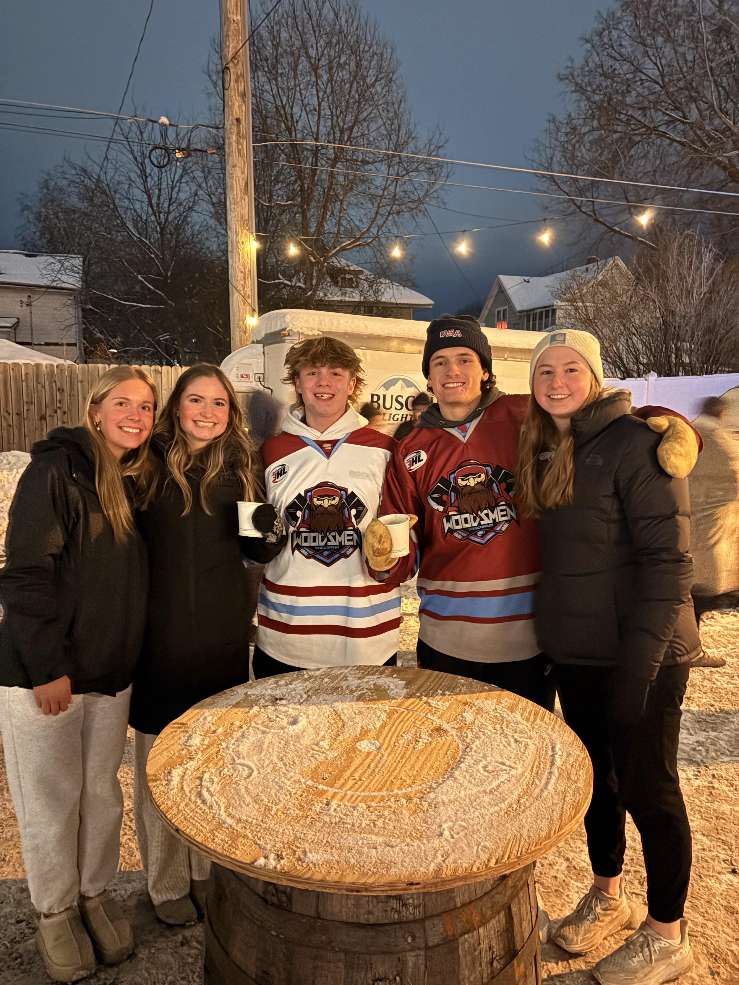 Five young adults, three women and two men, standing outdoors in winter by a large wooden table with a snow-covered surface, smiling at the camera. They are dressed warmly, some wearing hockey jerseys, hats, and gloves. There are snow-covered houses, trees, a wooden fence, and string lights illuminating the area in the background.