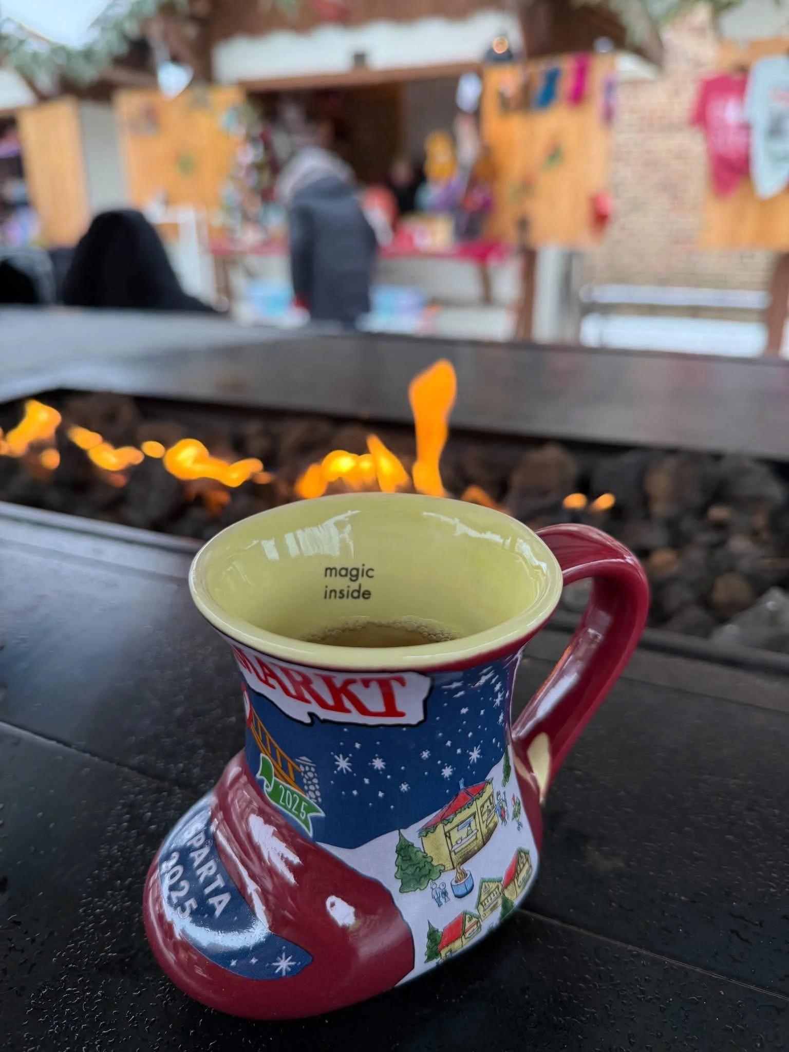 A holiday-themed ceramic mug with the words 'magic inside' visible at the top, placed on a black surface. The background shows an outdoor market or fair with blurred figures, tents, and colorful decorations.