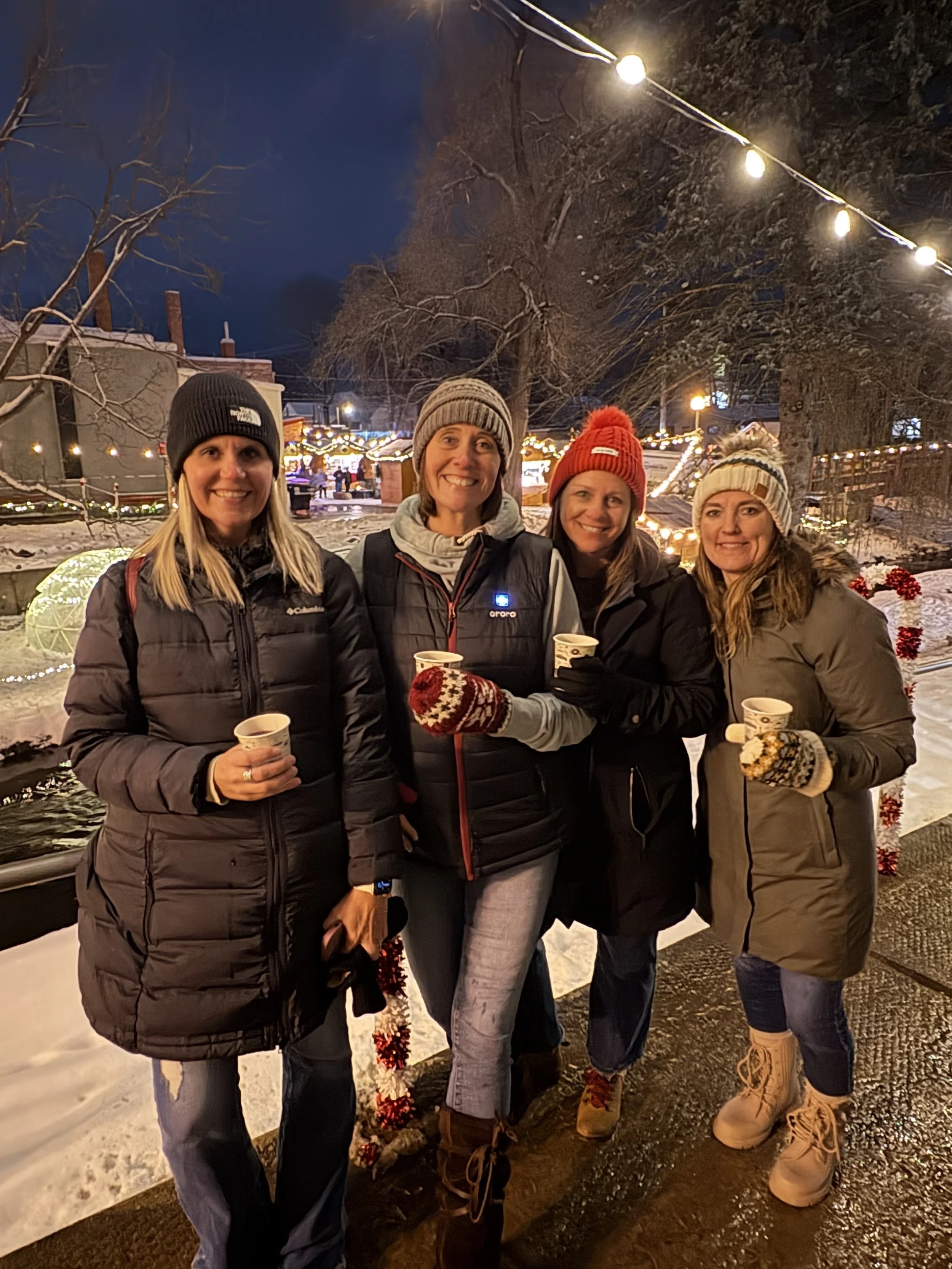 Four women dressed in winter clothing posing outdoors at night, holding cups, with Christmas lights and decorated trees in the background.