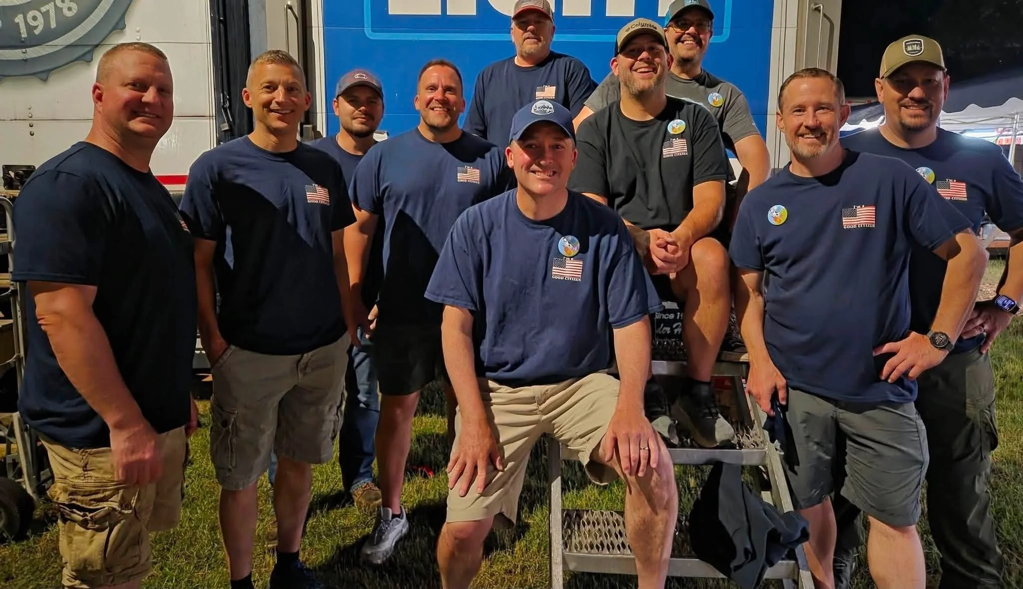 A group of ten men standing outdoors in front of a blue truck, wearing casual clothes and smiling for a photo, with some pins and American flag patches on their shirts.