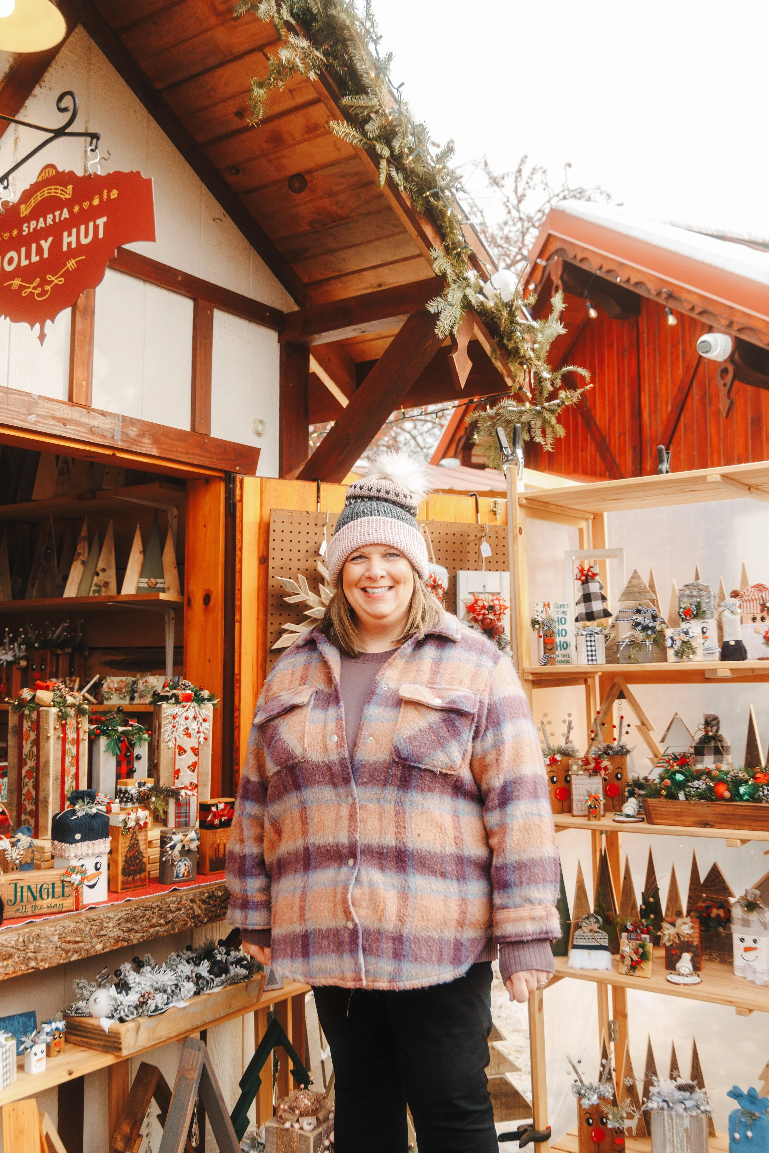 A woman stands in front of holiday craft decorations at a Christmas market stall, wearing a plaid coat and a knitted hat, smiling.