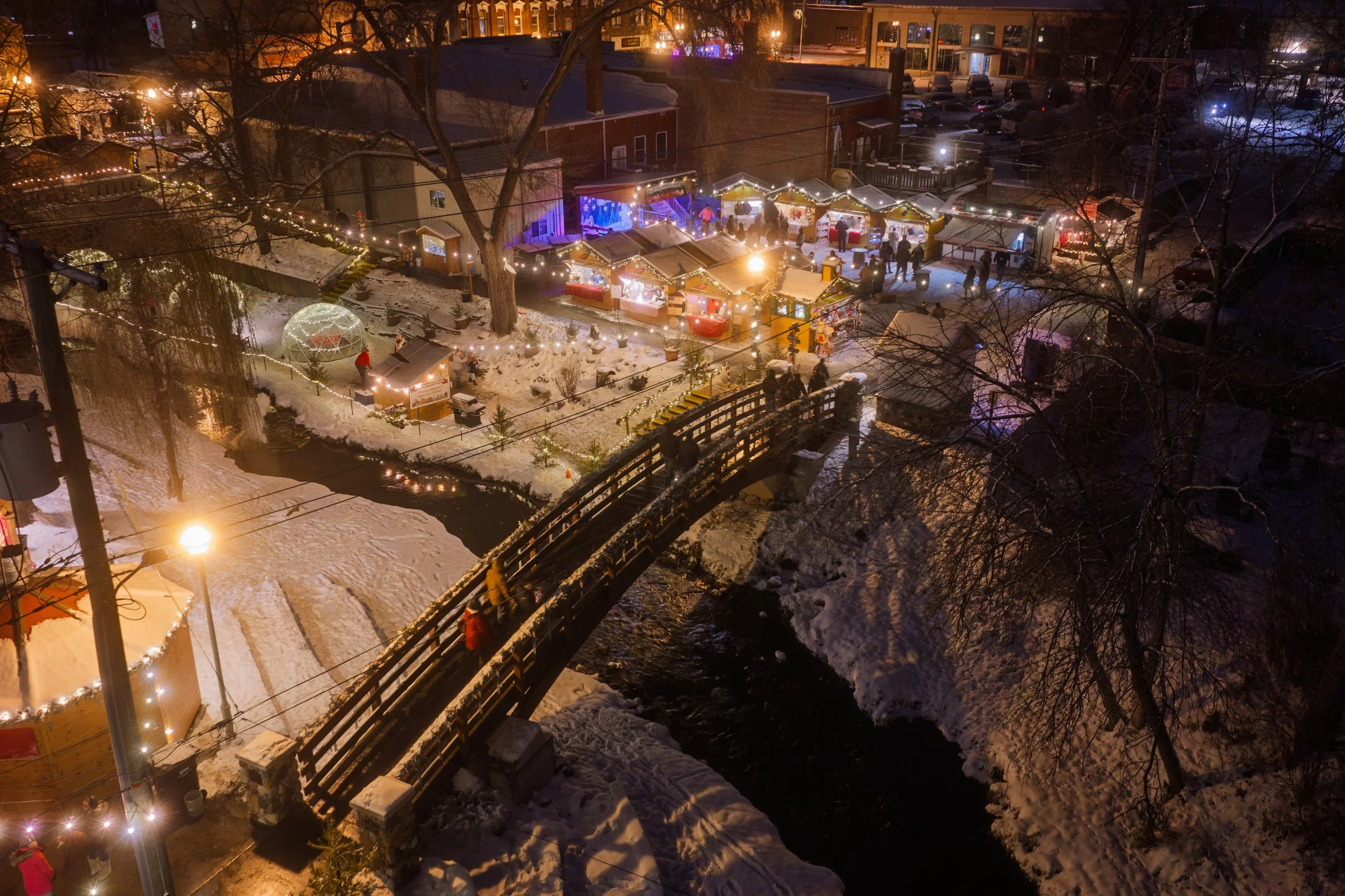 Nighttime scene of a Christmas market with decorated stalls, string lights, and people gathering, set in a snowy outdoor area with trees and a small stream.