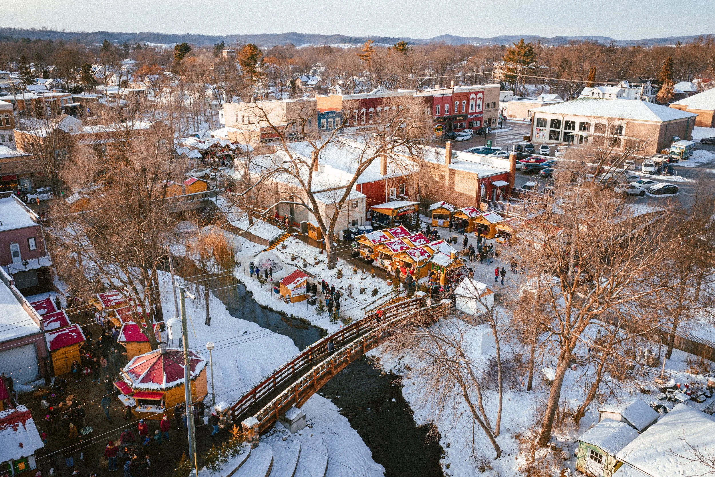 Aerial view of a snowy small town with a river, trees, buildings, and a street market with red and yellow tents.