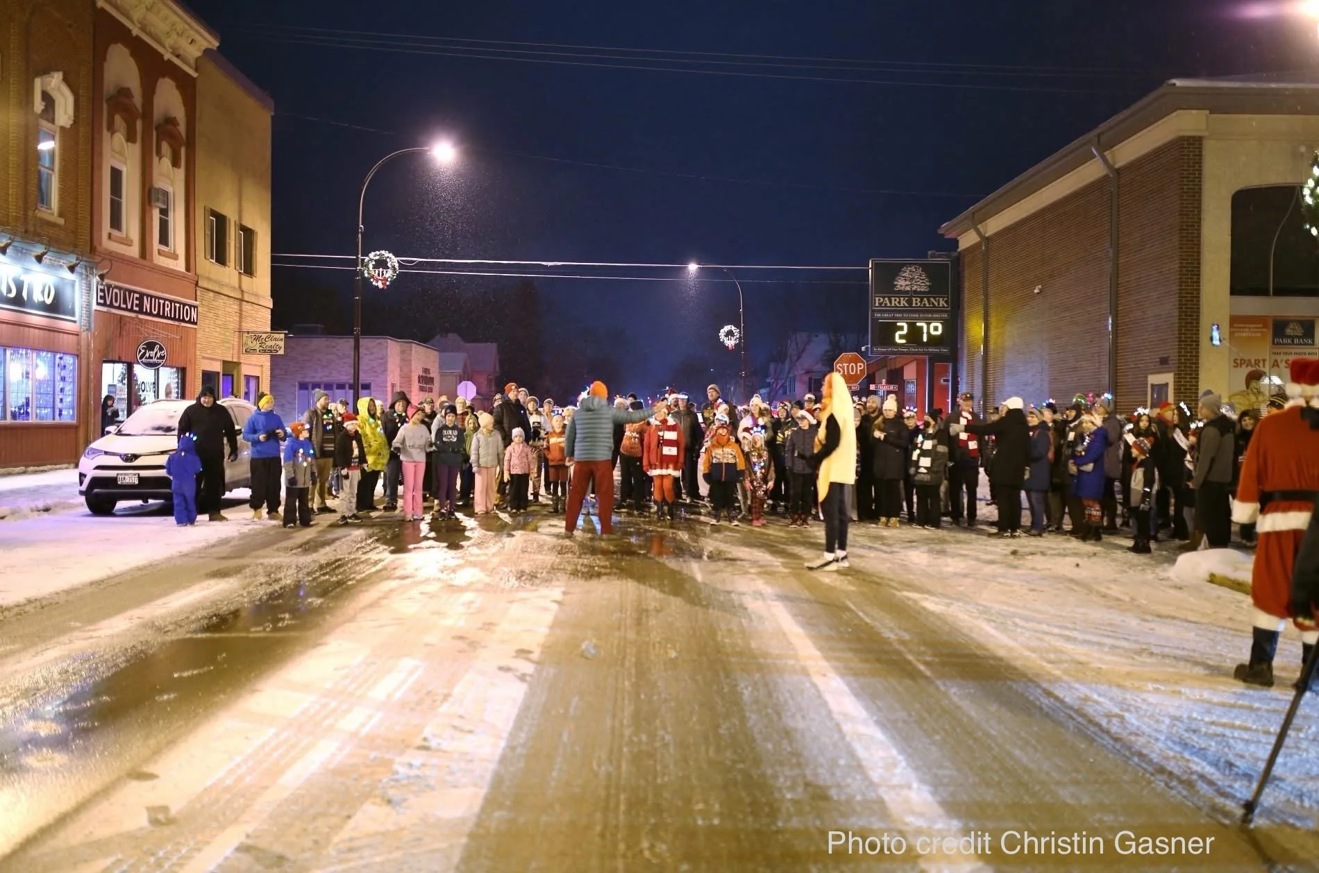 A large gathering of people in winter clothing on a snowy street at night, with festive decorations and streetlights, for a holiday or community event.