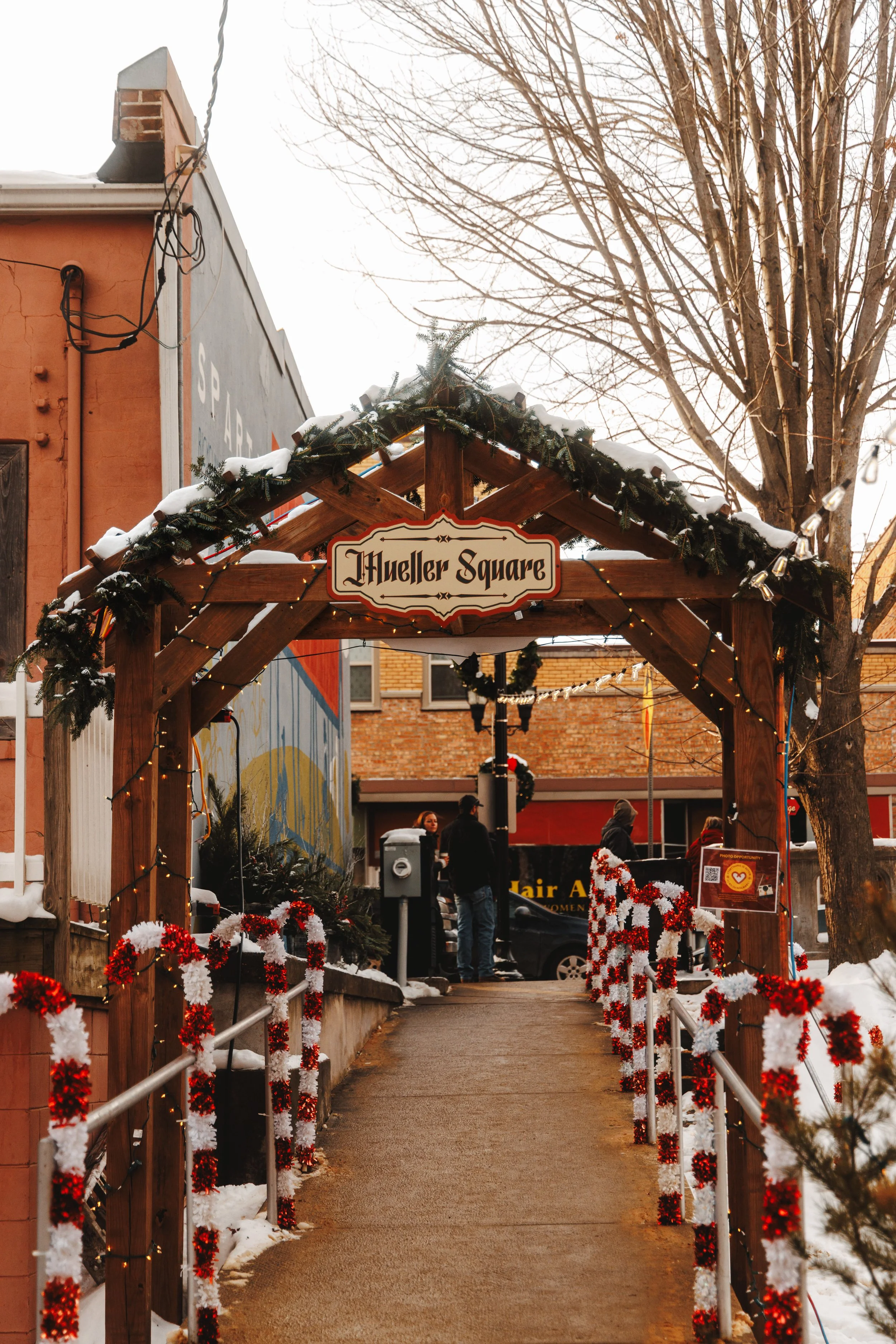 Wooden archway decorated with green garland, string lights, and red and white Christmas ornaments, with a sign that reads 'Mueller Square'. Snow is visible on parts of the decorations and ground, and people are seen walking in the background.